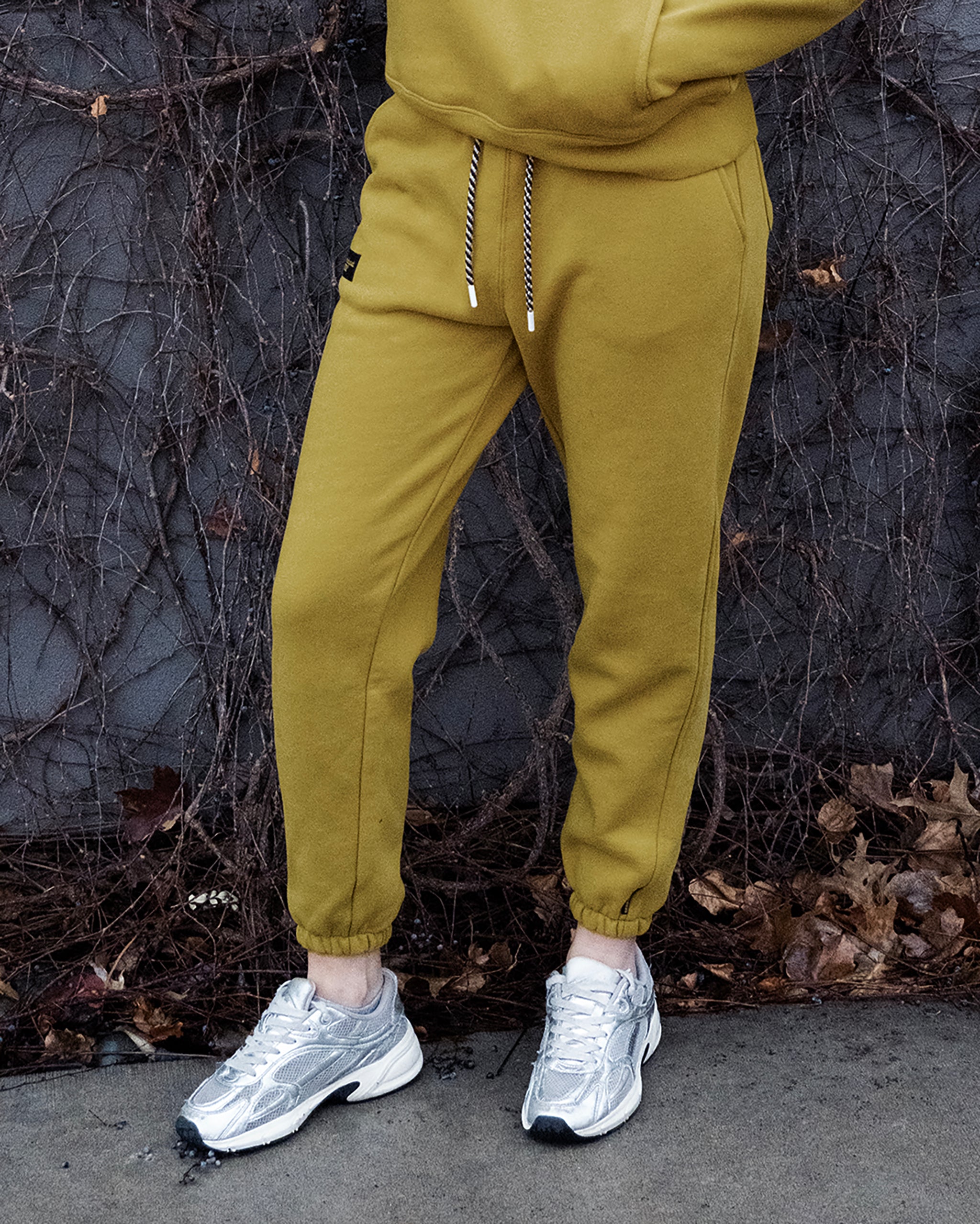 A person, shown from the shoulders down, wears BEARDEDGOAT's NEW — Overnight™ Classic Jogger with a high-rise waistband and matching sweatshirt, paired with shiny silver sneakers, while standing before a wall lined with bare vines and dried leaves.