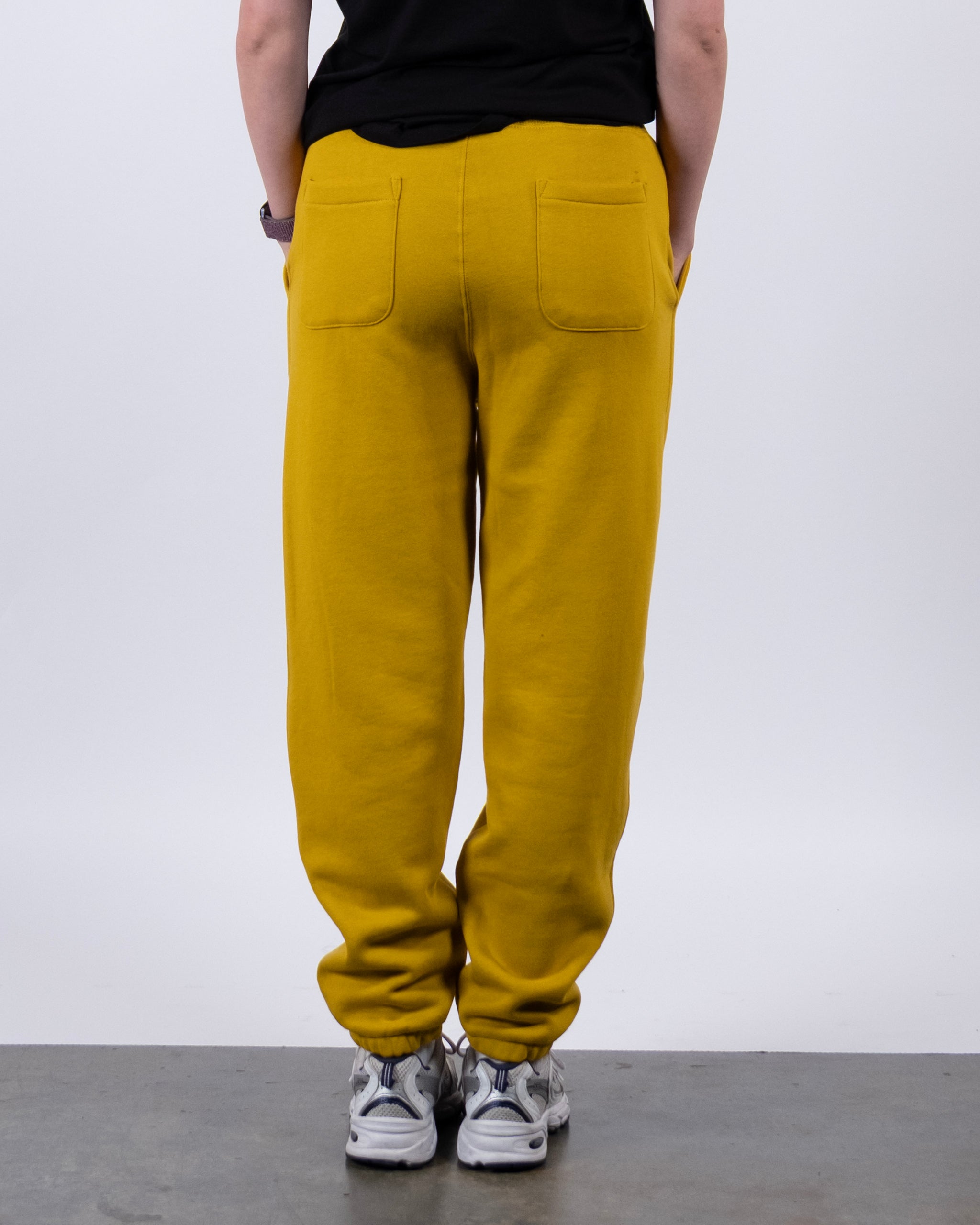A person faces away from the camera, wearing BEARDEDGOAT's NEW — Overnight™ Classic Jogger in mustard yellow with patch pockets and a high-rise waistband, paired with white sneakers against a plain, light background.