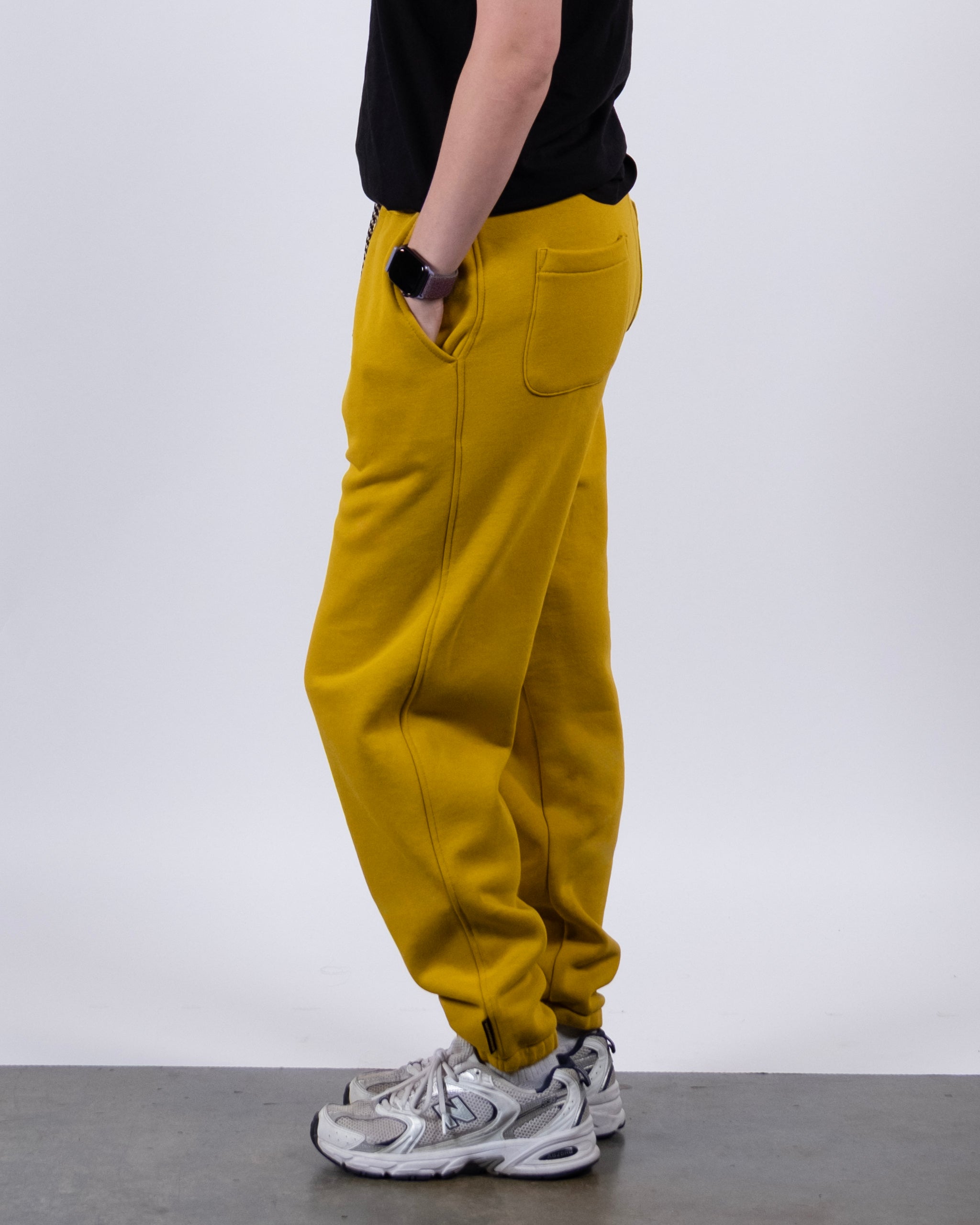 A person wearing BEARDEDGOAT NEW — Overnight™ Classic Jogger in mustard yellow, a black t-shirt, white sneakers, and a smartwatch stands sideways with hands in pockets against a plain white background.