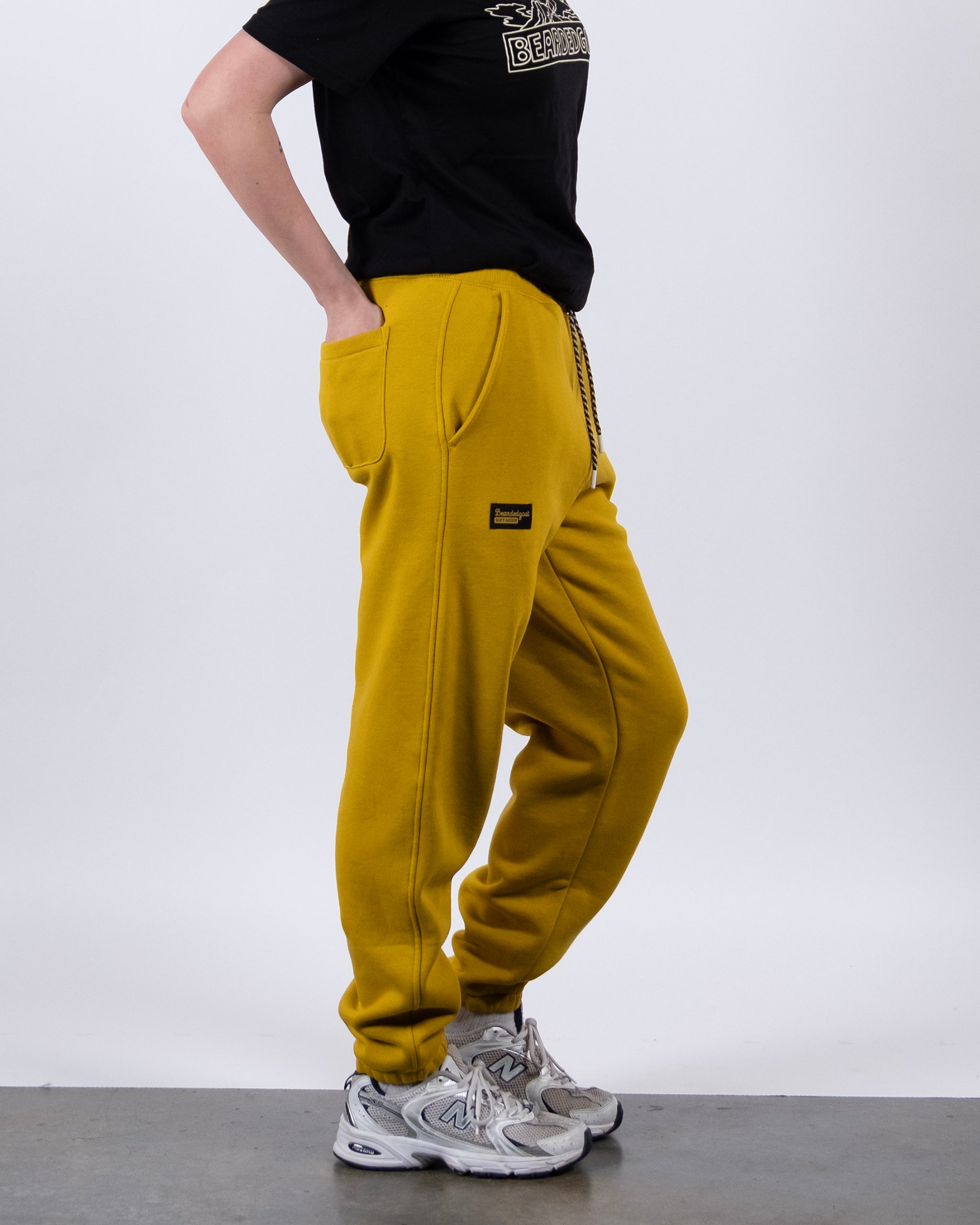 A person stands sideways with hands in pockets, wearing a black tee, BEARDEDGOAT NEW — Overnight™ Classic Jogger in bright yellow featuring a high-rise waistband, and light gray sneakers, all set against a plain white background.