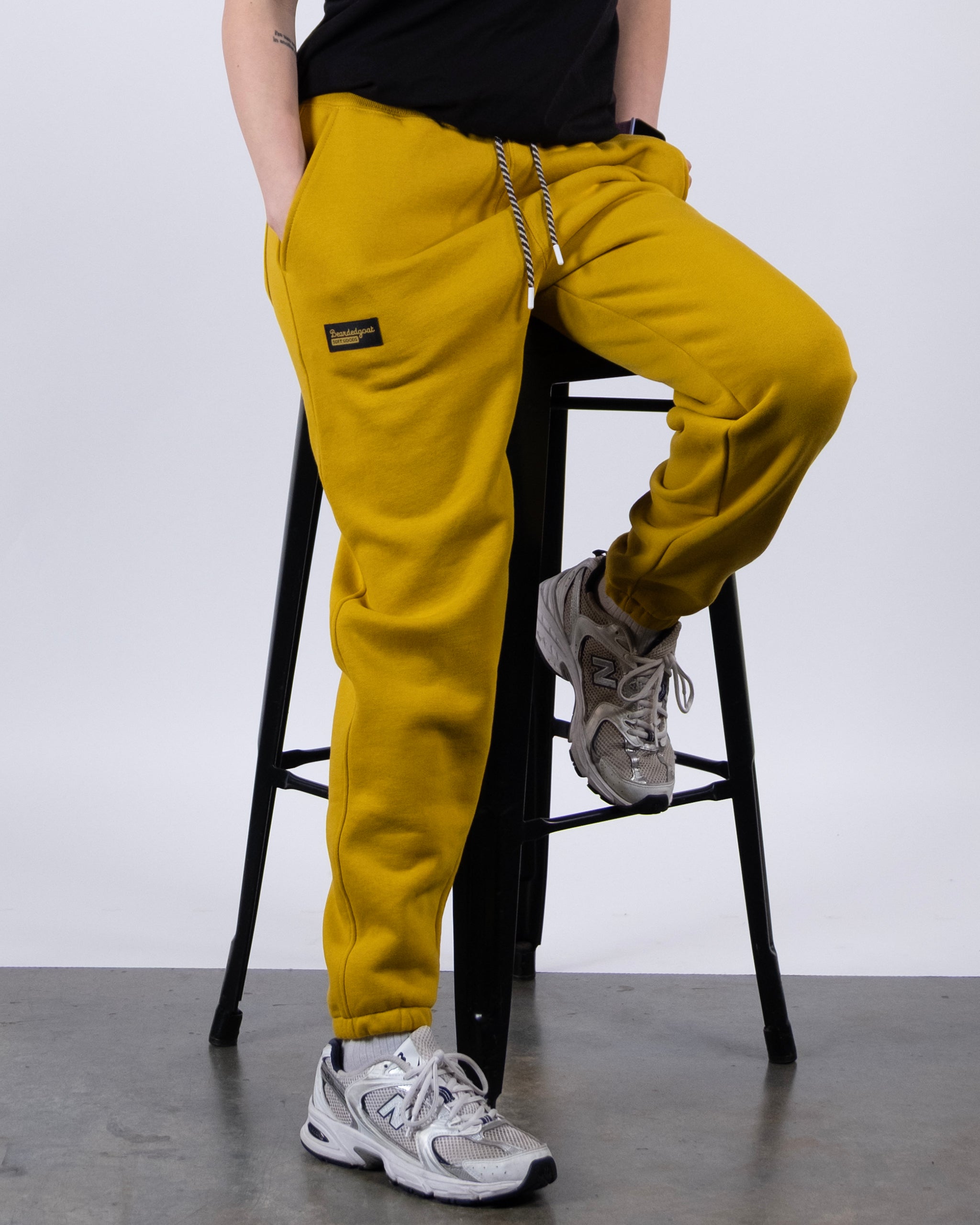 A person sits on a black stool against a white background, wearing BEARDEDGOAT NEW — Overnight™ Classic Jogger sweatpants in bright yellow and grey New Balance sneakers. Only the lower half of their body is visible.