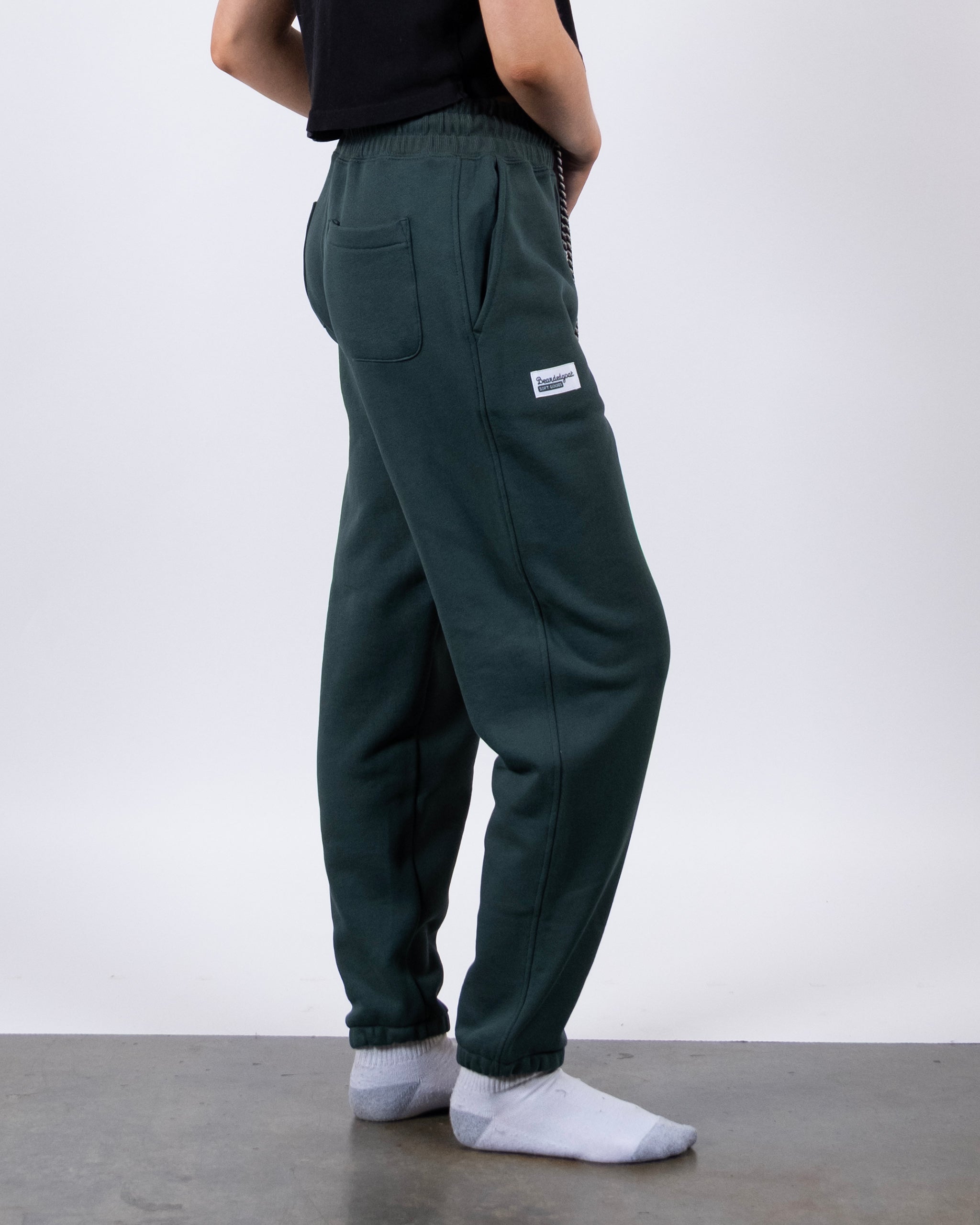 A person stands on a gray floor, wearing BEARDEDGOAT's NEW — Overnight™ Classic Jogger in dark green heavyweight cotton fleece, paired with a black shirt and white socks, all set against a plain white background.