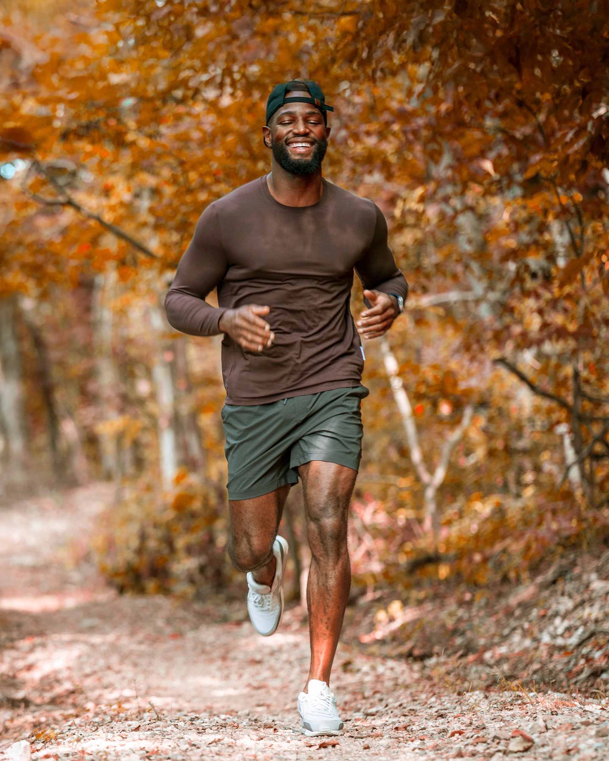 A man wearing the BEARDEDGOAT Men’s EverZen™ Long Sleeve jogs and smiles along a dirt trail through an autumn forest, paired with shorts.