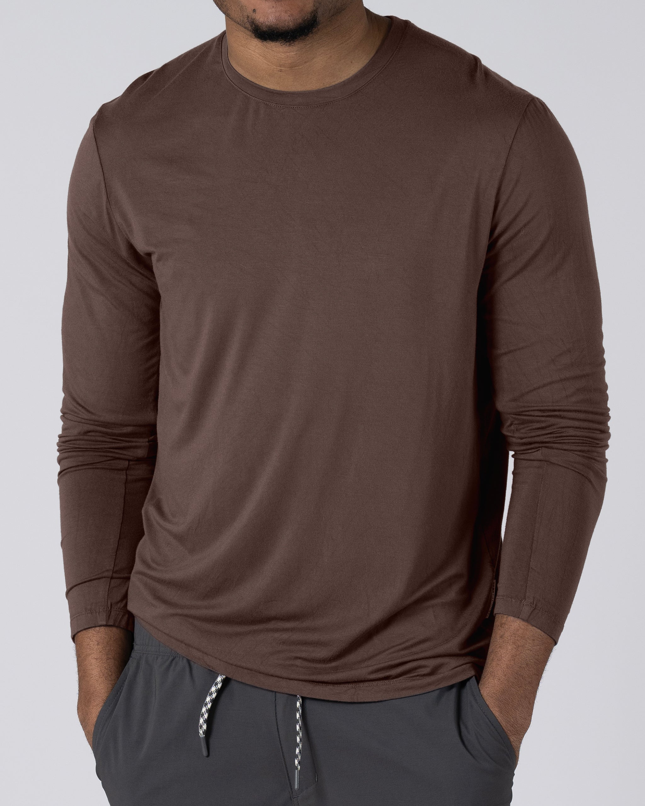 A man wears the BEARDEDGOAT Men’s EverZen™ Long Sleeve and gray drawstring pants, standing with hands in his pockets against a plain light background. His face is partially out of view.