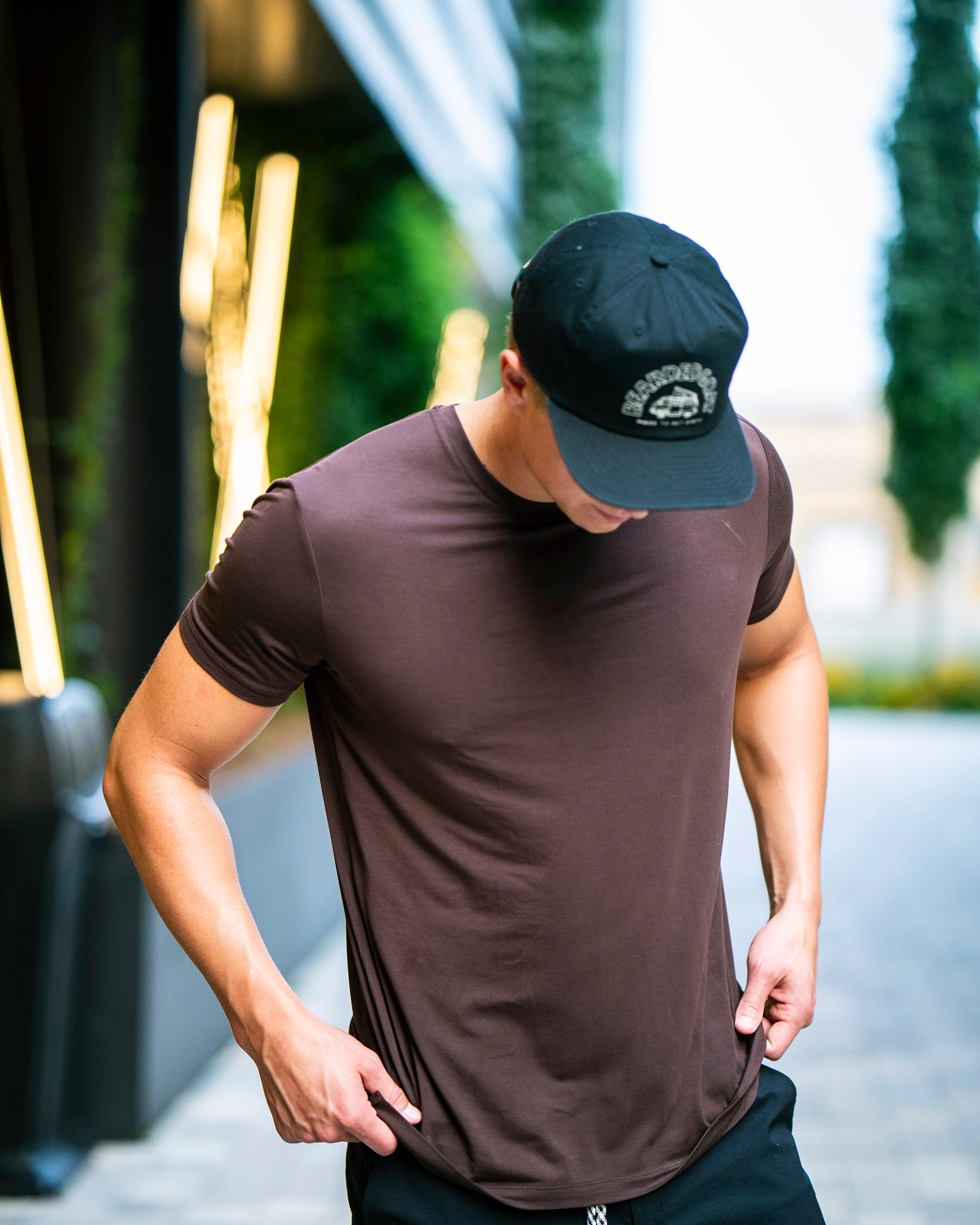 A man in a black cap and a BEARDEDGOAT Men’s EverZen™ Short Sleeve adjusts his shirt while standing outside on a city street, with blurred lights and greenery in the background.
