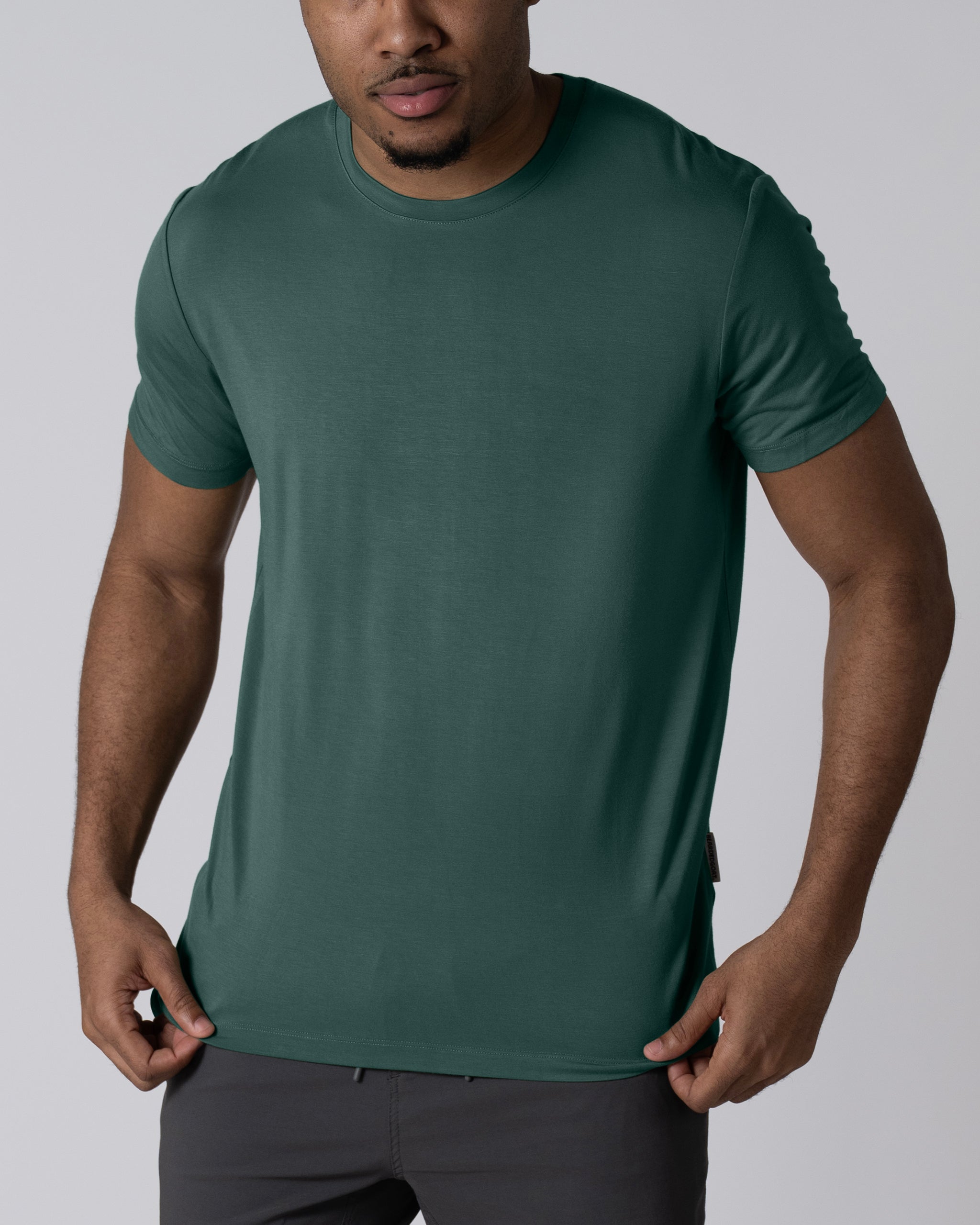 A man wearing the BEARDEDGOAT Men’s EverZen™ Short Sleeve in dark green and dark pants stands against a light gray background, holding the sides of his shirt. His face is partially visible. The bamboo blend fabric offers performance comfort.