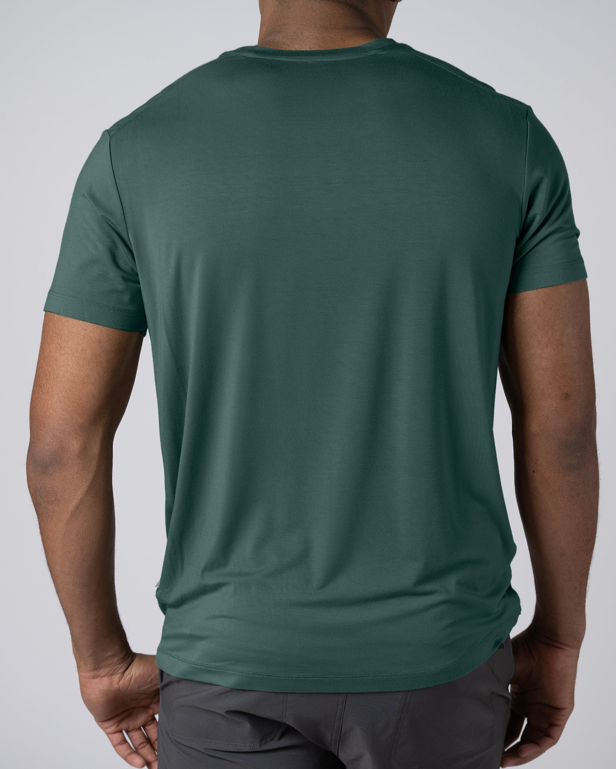 A person is shown from the back wearing a BEARDEDGOAT Men’s EverZen™ Short Sleeve, a dark green bamboo-blend performance t-shirt, paired with dark gray pants, standing against a plain light gray background.