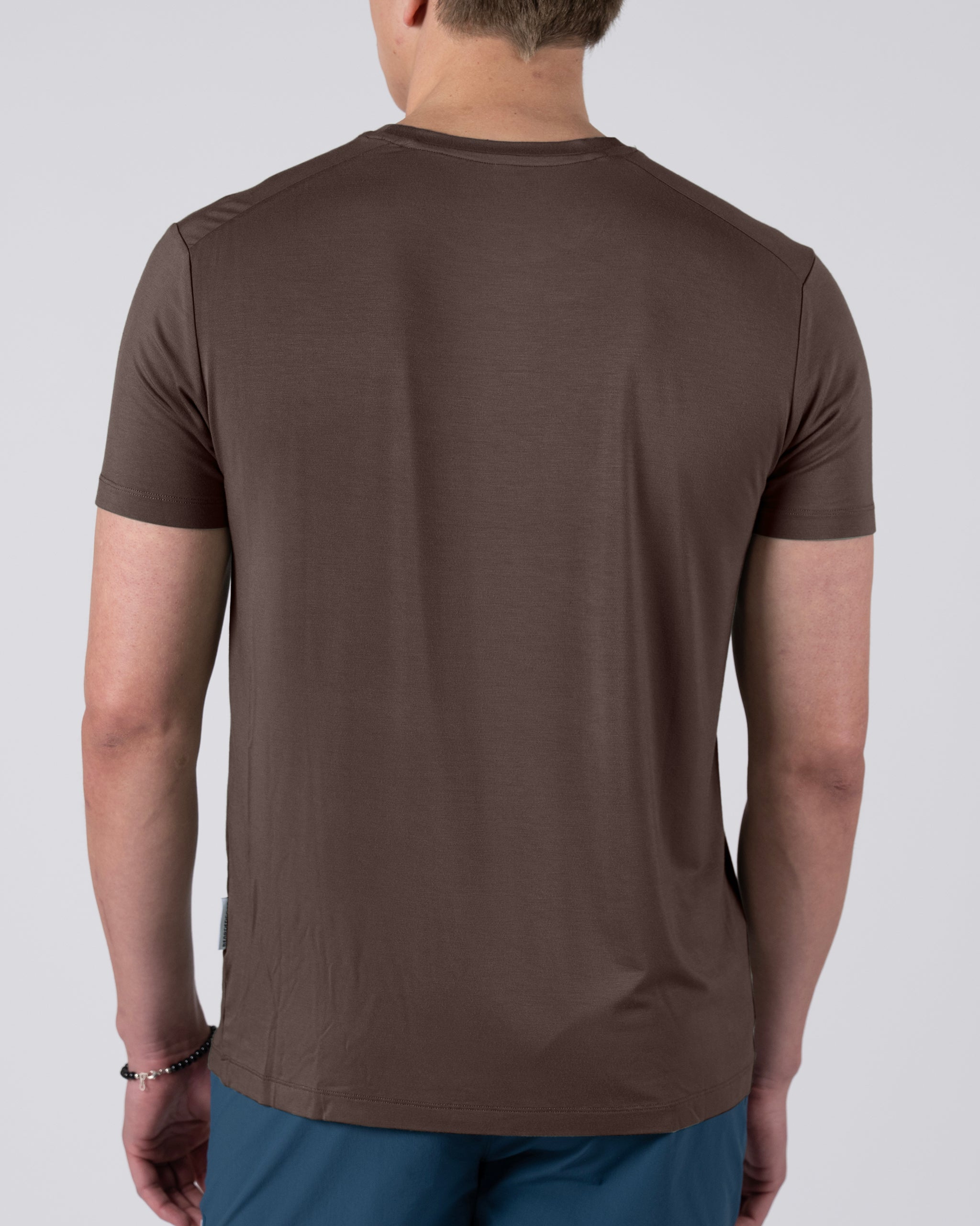A man is seen from the back wearing a BEARDEDGOAT Men’s EverZen™ Short Sleeve brown t-shirt and blue pants, standing with relaxed arms against a plain light background.