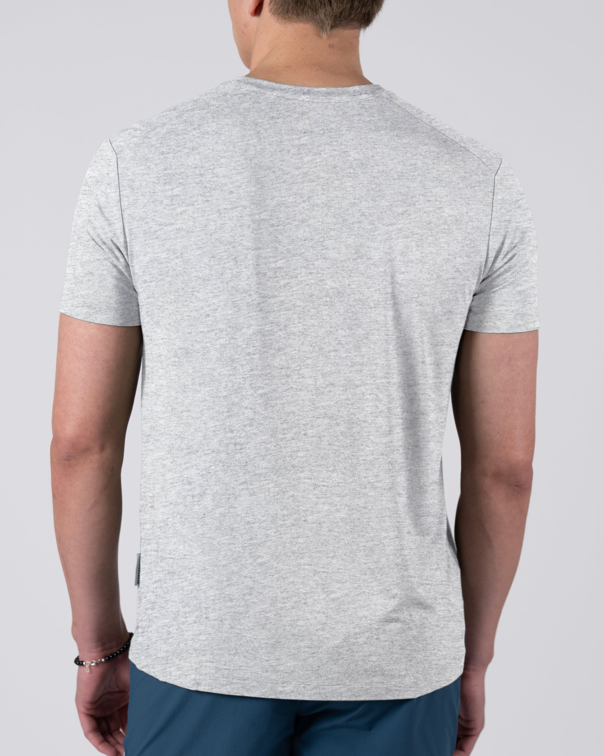 A person is seen from behind wearing a BEARDEDGOAT Men’s EverZen™ Short Sleeve t-shirt in light grey and teal pants, standing against a plain light background.