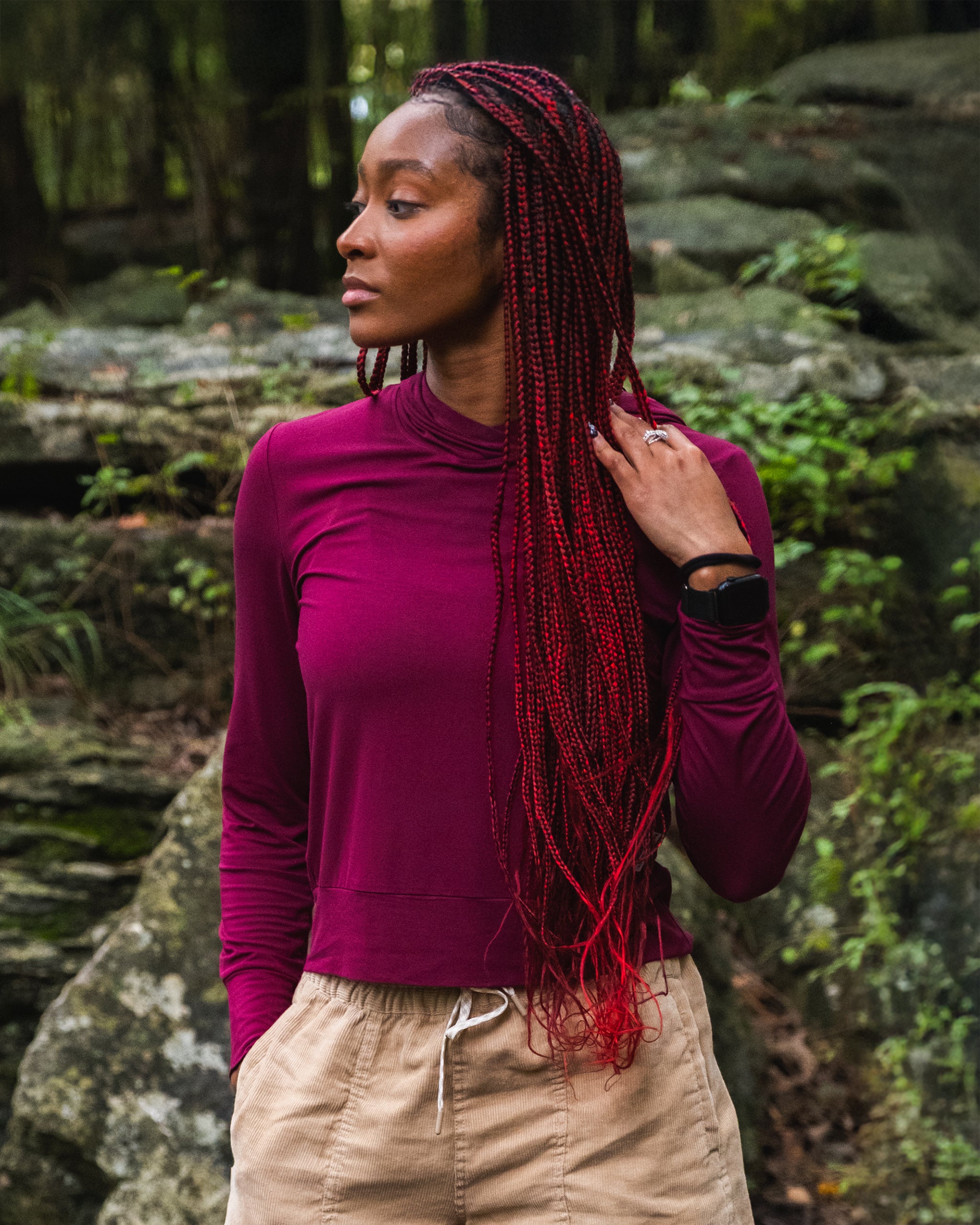 A woman with long red braids, wearing a burgundy BEARDEDGOAT Women’s EverZen™ Bamboo Hoodie and tan pants, stands outdoors before rocks and greenery, looking to the side while holding her hair.