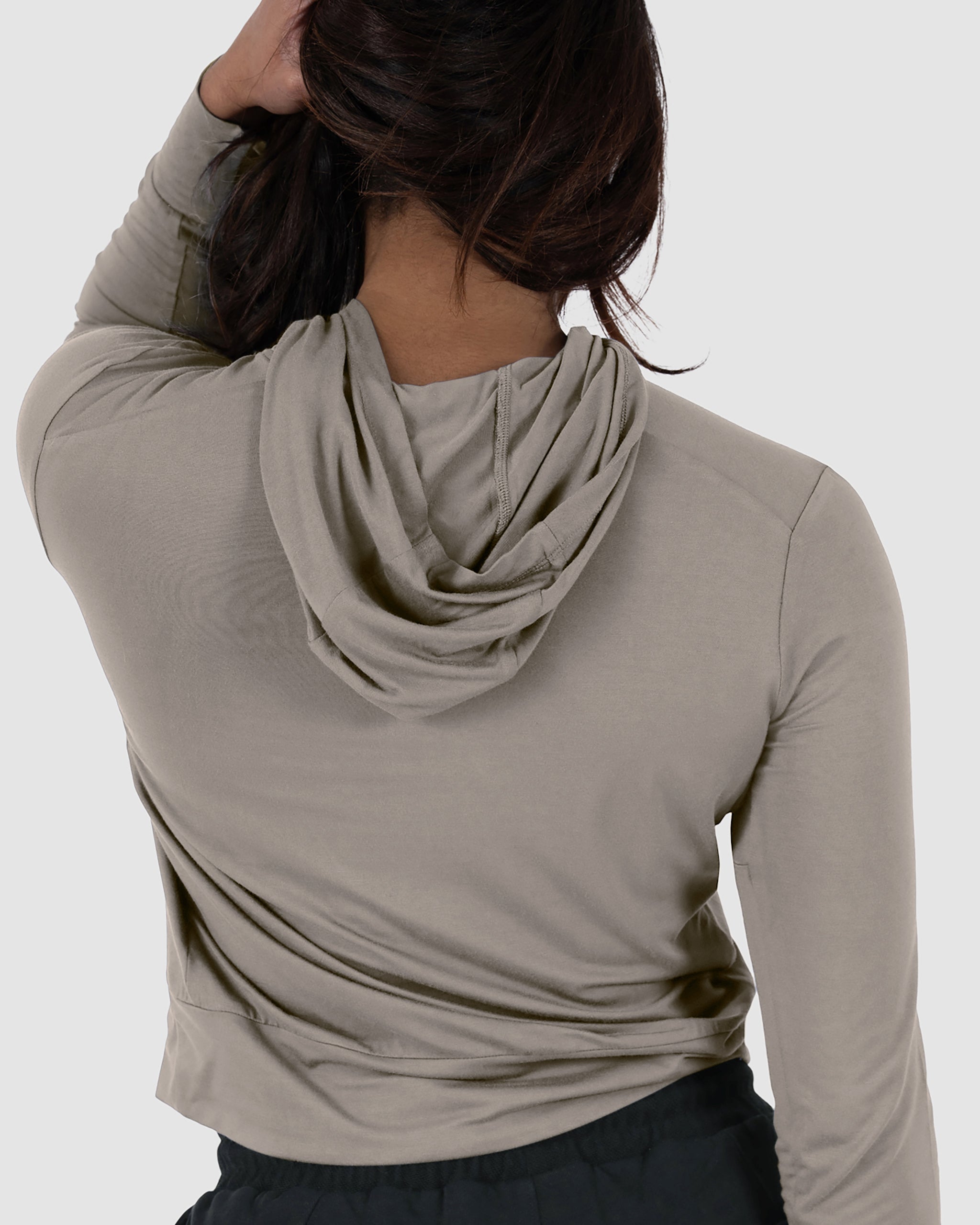 A person with long dark hair, seen from behind, wears a light beige BEARDEDGOAT Women’s EverZen™ Bamboo Hoodie and black pants with the hood down against a plain light gray background.