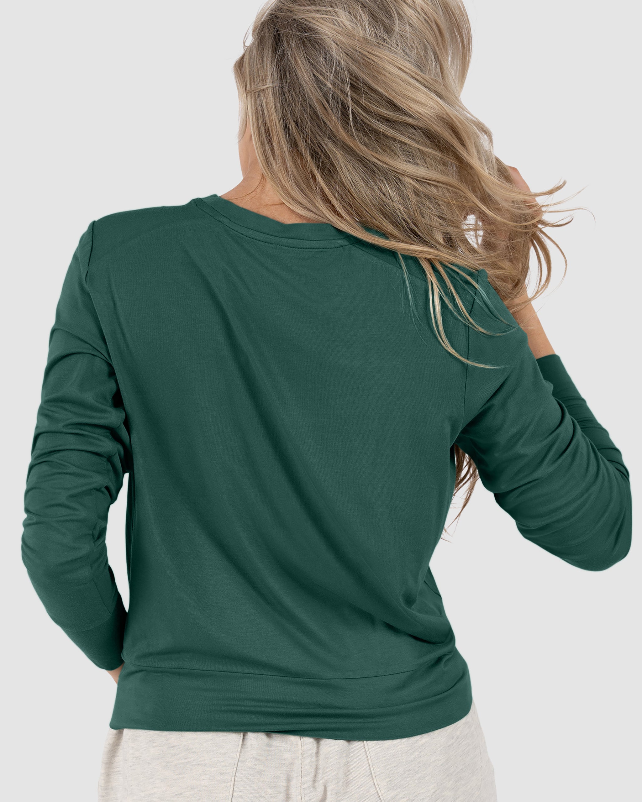 A woman with long, wavy blonde hair is seen from behind, wearing a dark green BEARDEDGOAT Women’s EverZen™ Bamboo Long Sleeve and light-colored pants against a plain light background.