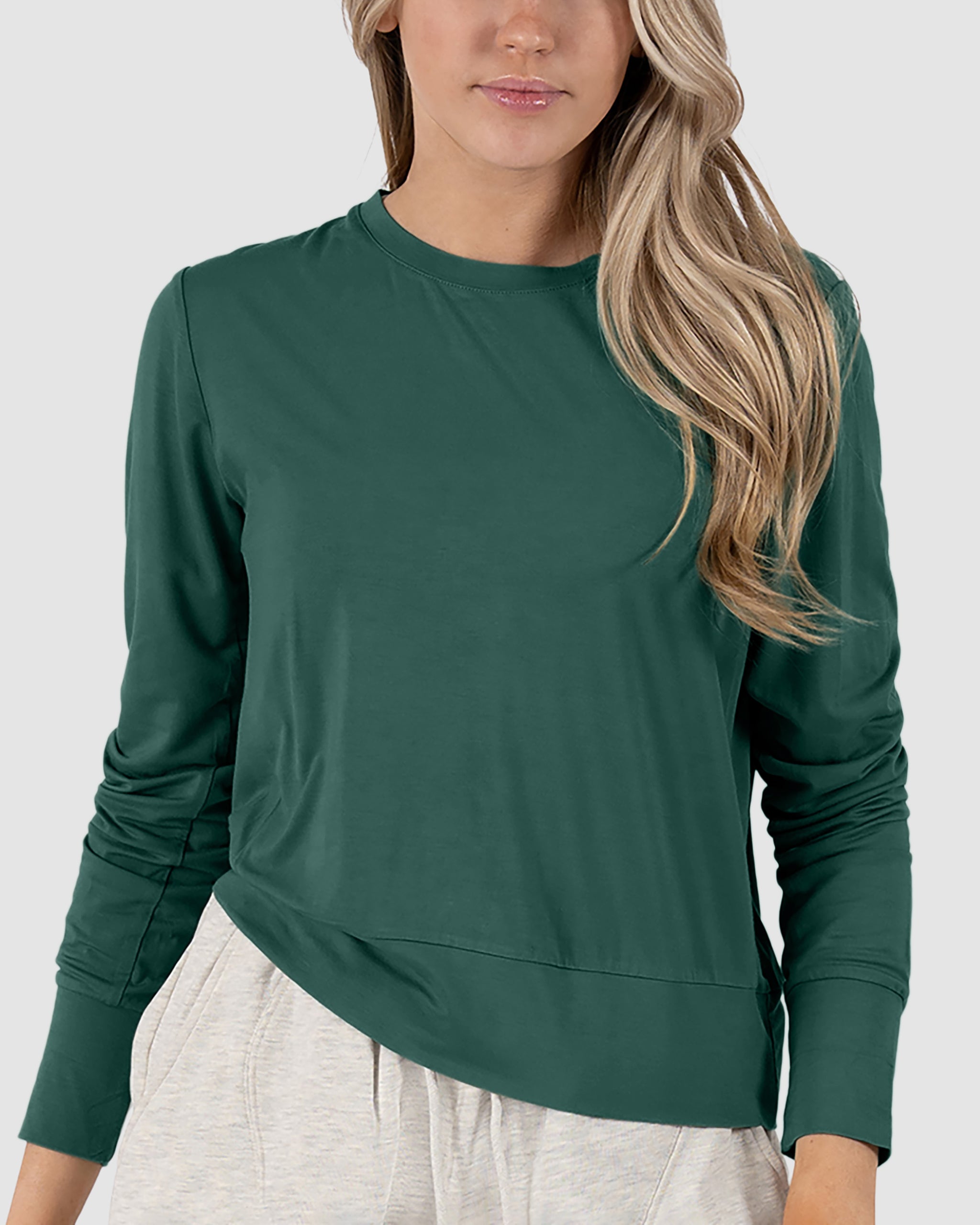 A woman with long blonde hair wears a BEARDEDGOAT Women’s EverZen™ Bamboo Long Sleeve in Cypress and light-colored pants, standing against a plain light gray background.