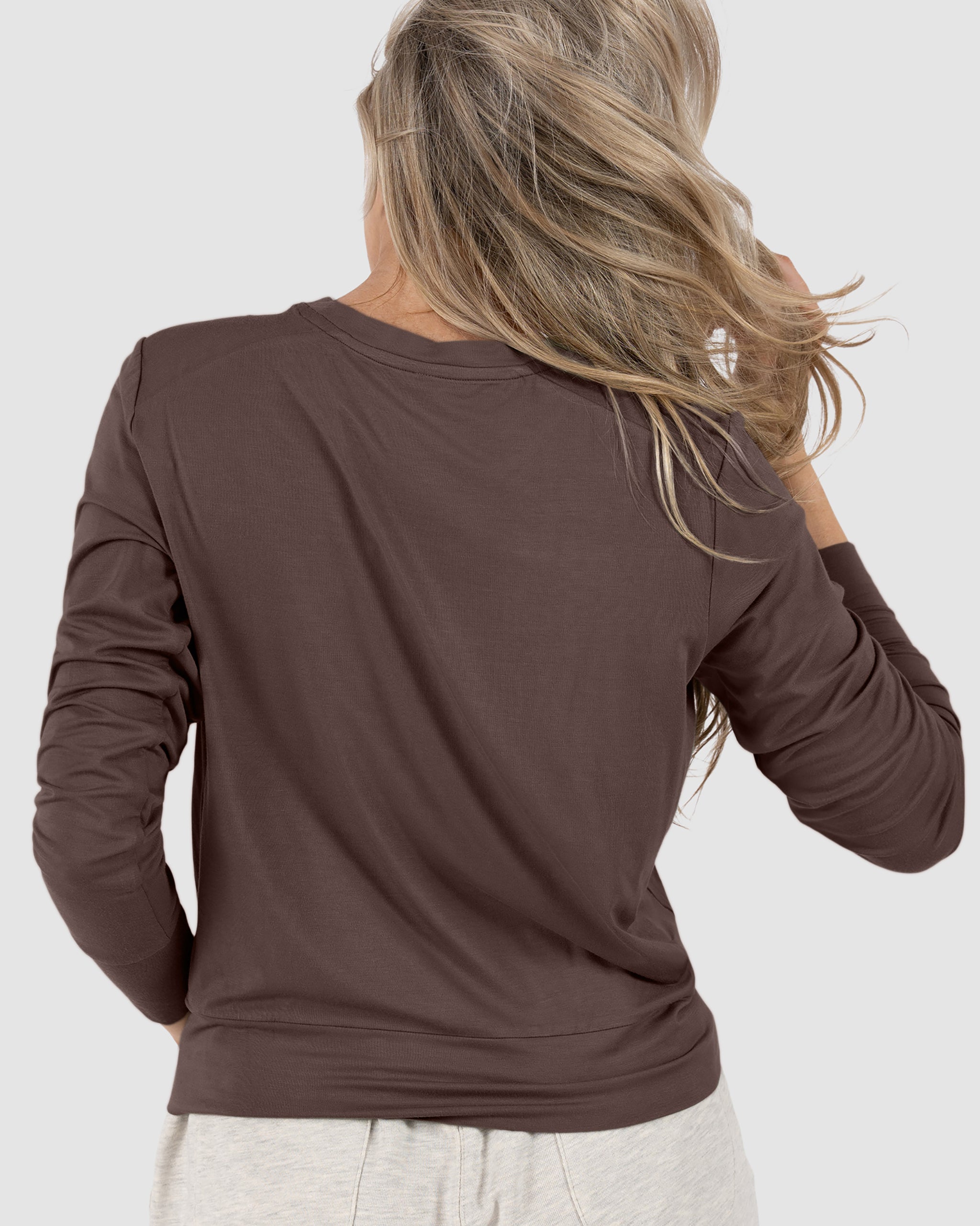 A woman with long, wavy blonde hair is seen from behind, wearing the BEARDEDGOAT Women’s EverZen™ Bamboo Long Sleeve in brown and light bamboo-blend pants, set against a plain light gray background.