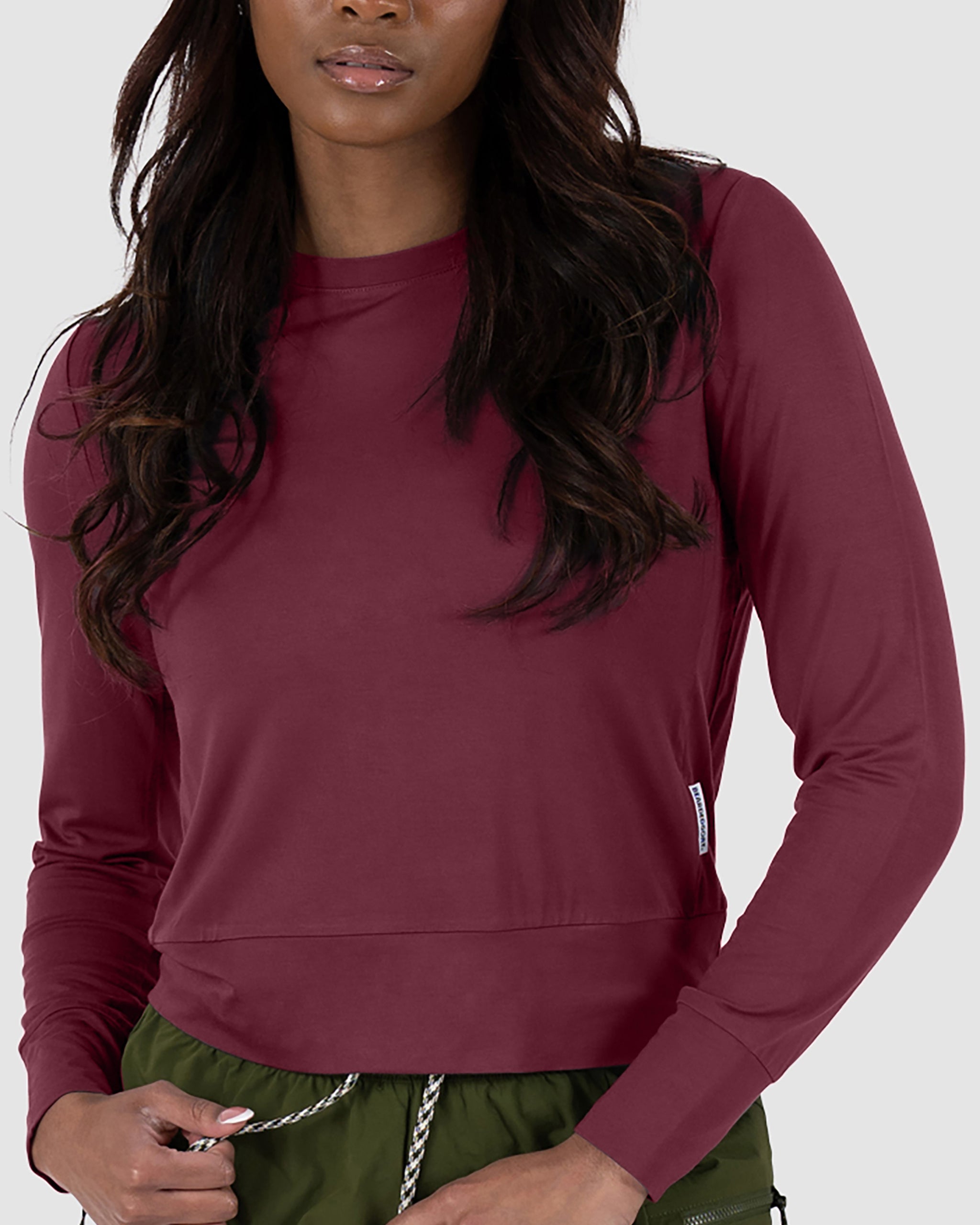 A woman wears the BEARDEDGOAT Women’s EverZen™ Bamboo Long Sleeve in Rustic Ember, paired with olive green drawstring pants, standing against a light gray background.