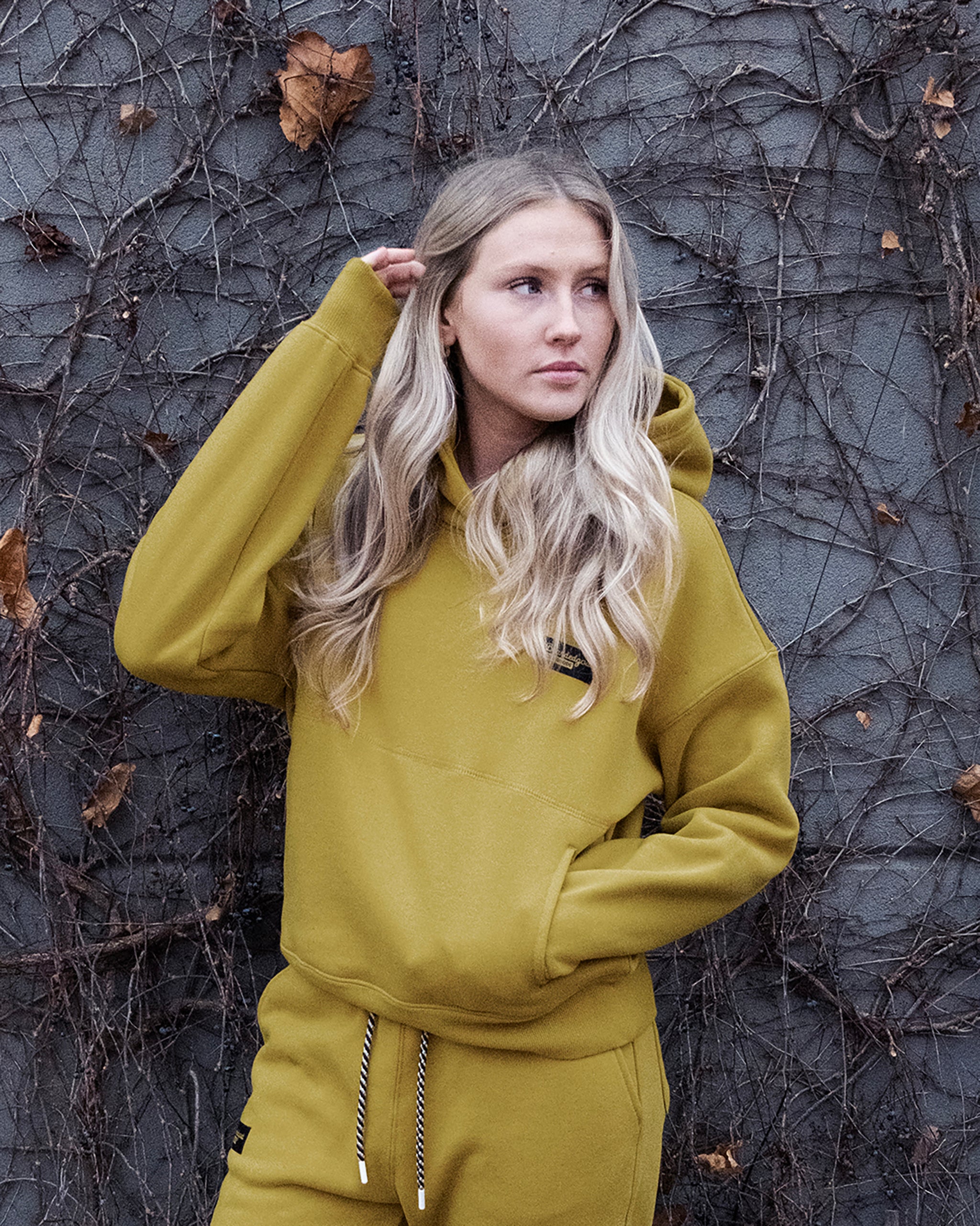 A young woman with long blonde hair wears a mustard yellow beardedgoat Women's Overnight™ Hoodie and sweatpants made from heavyweight cotton fleece, standing before a gray vine-covered wall and looking to the side with one hand in her hair.