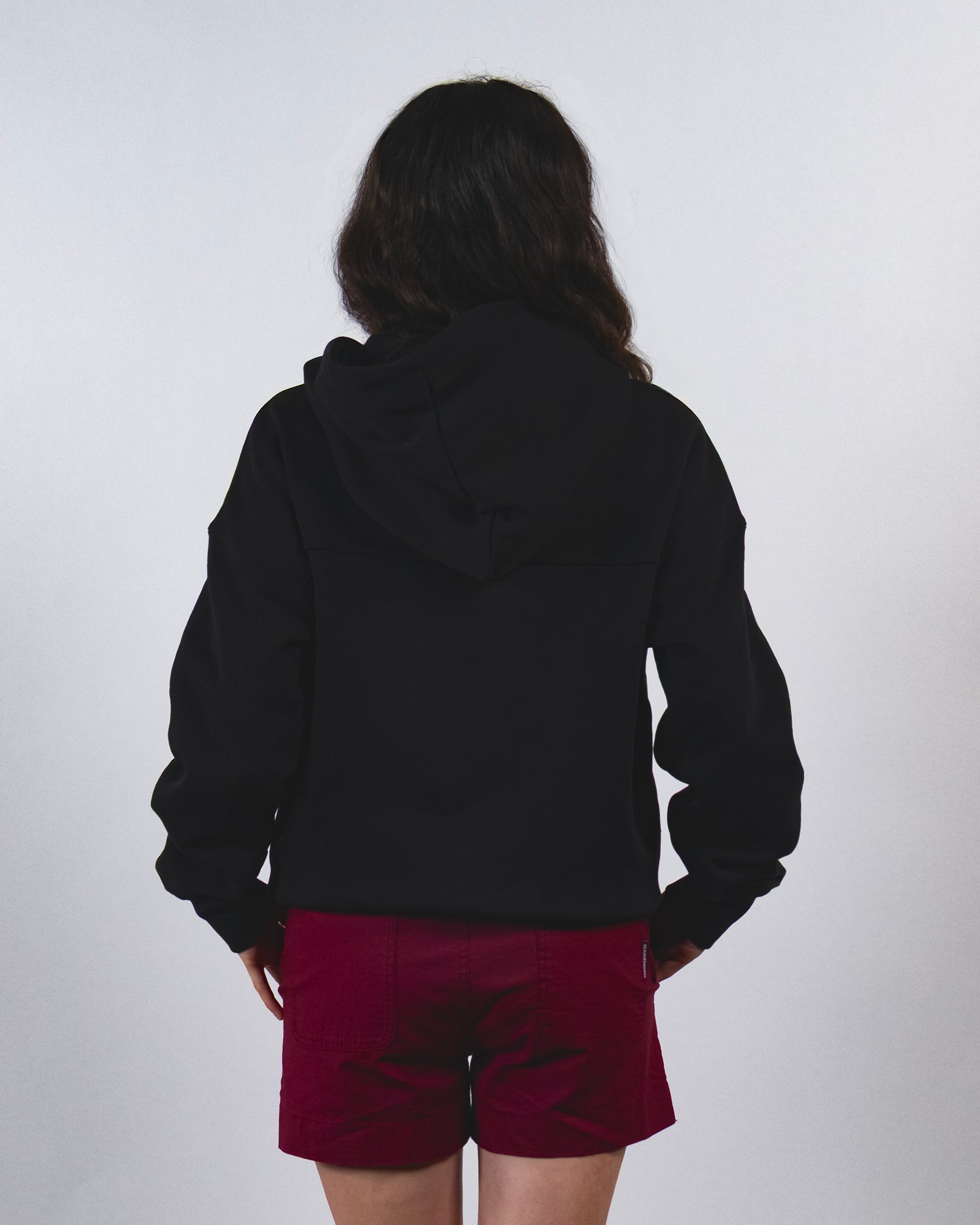 A person with long dark hair, wearing a black Women's Overnight™ Hoodie by beardedgoat paired with red shorts, stands facing away from the camera against a plain light-colored background.