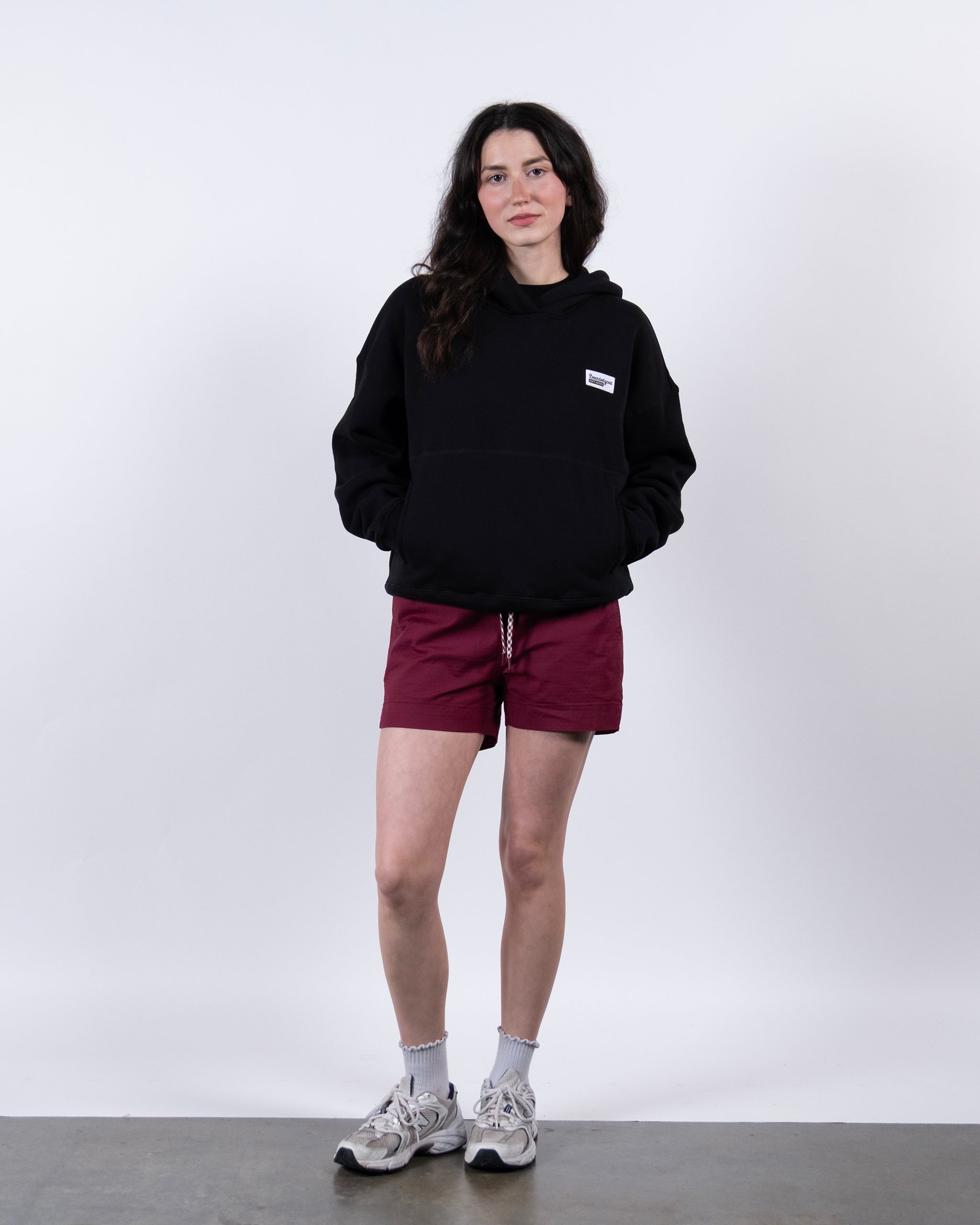 A woman with long dark hair stands against a white background, wearing a beardedgoat Women's Overnight™ Hoodie, maroon shorts, white socks, and gray sneakers. Her hands are in her hoodie pockets and she has a neutral expression.