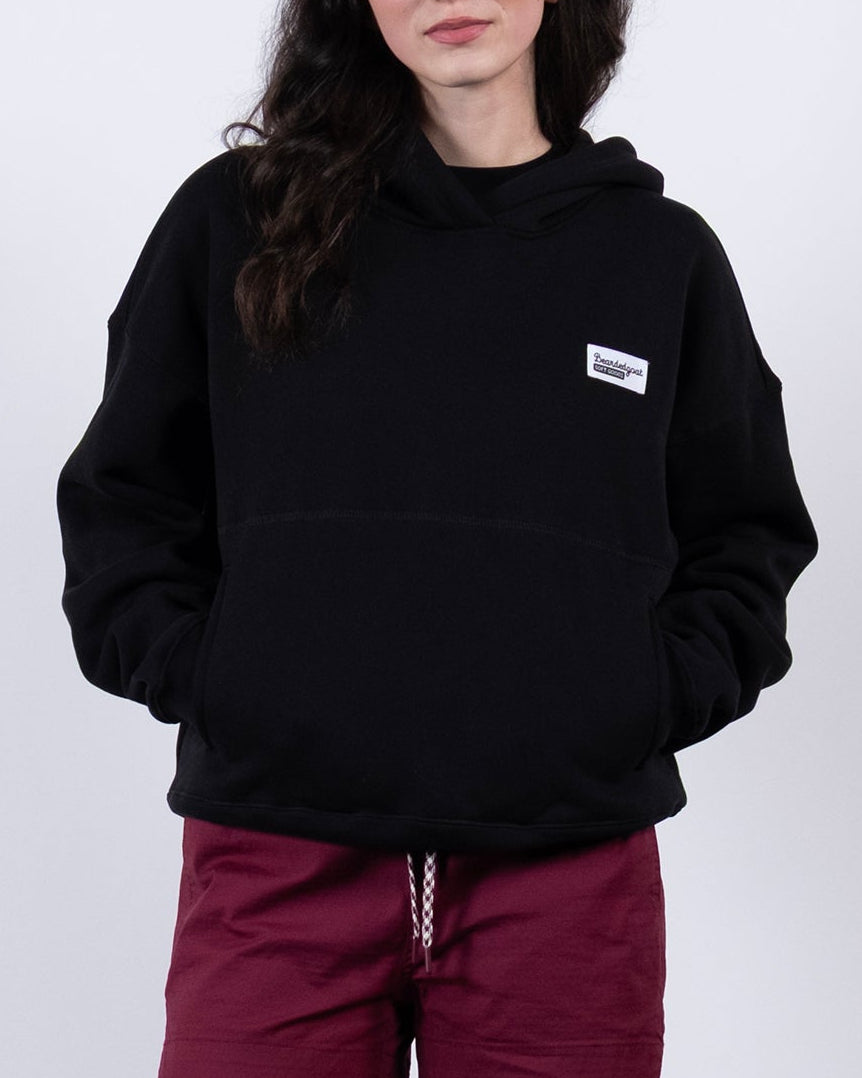 A woman with long dark hair stands against a white background, wearing a beardedgoat Women's Overnight™ Hoodie, maroon shorts, white socks, and gray sneakers. Her hands are in her hoodie pockets and she has a neutral expression.