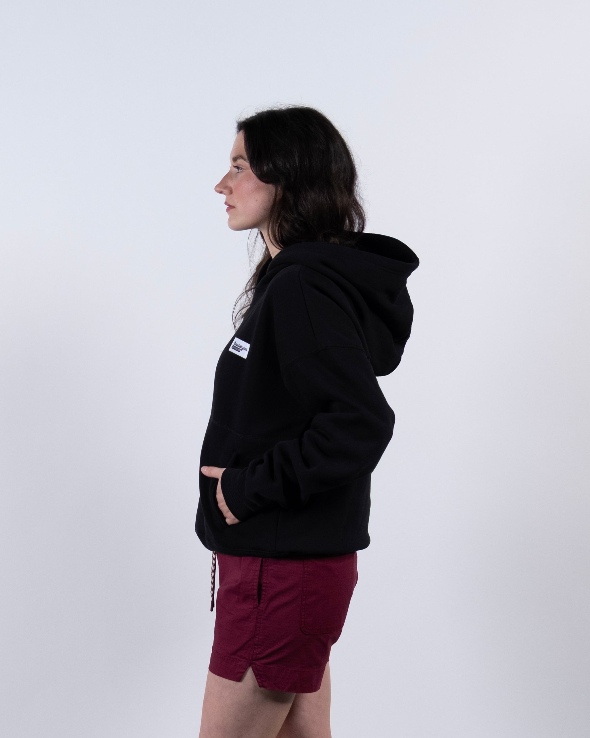 A woman with long dark hair stands in profile against a white background, hands in her pockets, wearing the beardedgoat Women's Overnight™ Hoodie made from premium cotton fleece and maroon shorts.