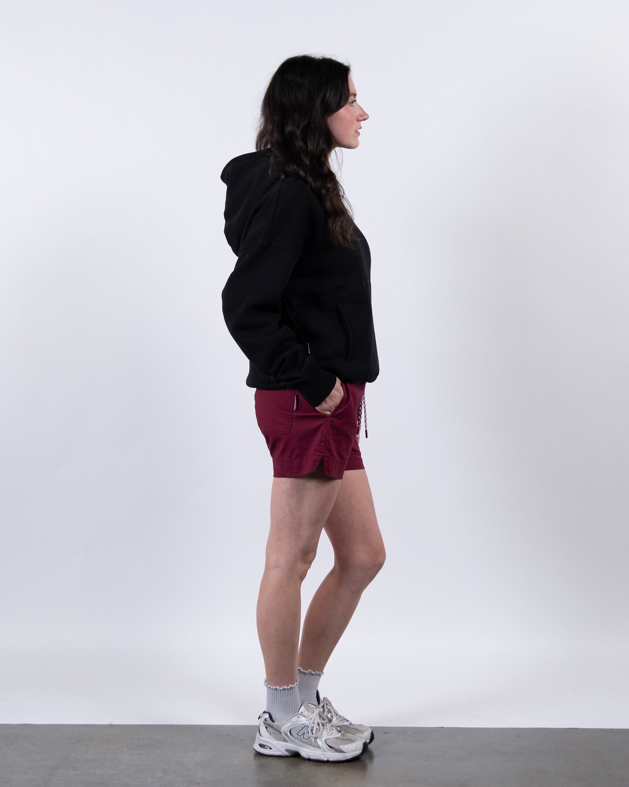 A woman with long dark hair stands in profile against a white background, wearing the beardedgoat Women's Overnight™ Hoodie, maroon shorts, gray socks, and white sneakers. Her hands are in her pockets as she looks to the left.