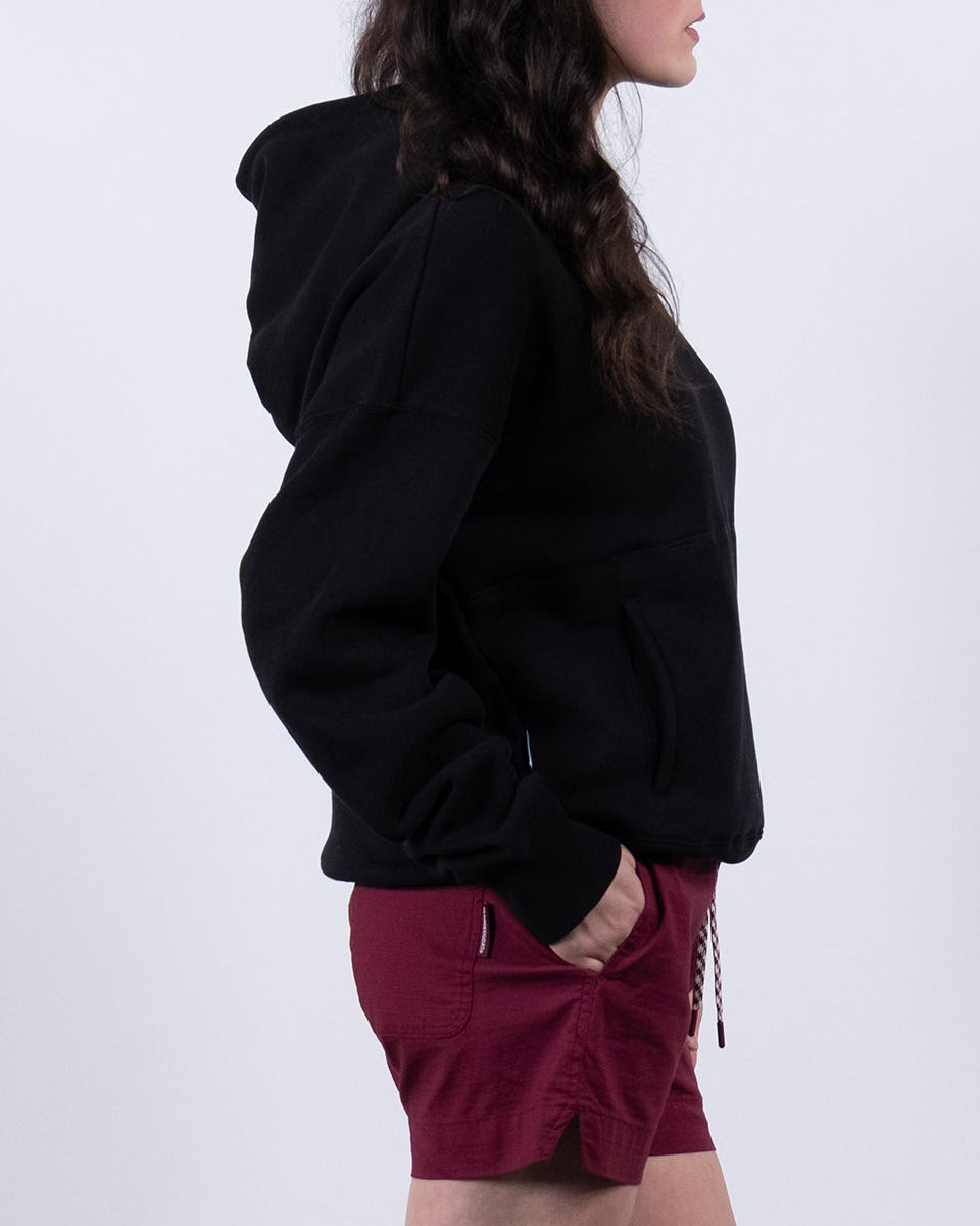 A woman with long dark hair stands in profile against a white background, wearing the beardedgoat Women's Overnight™ Hoodie, maroon shorts, gray socks, and white sneakers. Her hands are in her pockets as she looks to the left.