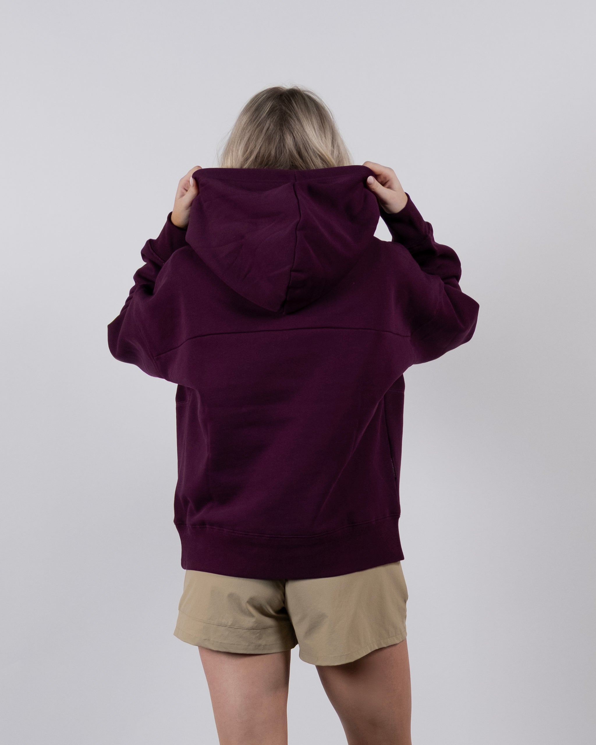 A person faces away from the camera, wearing the beardedgoat Women's Overnight™ Hoodie in maroon with the hood up and beige shorts, standing against a plain light gray background.