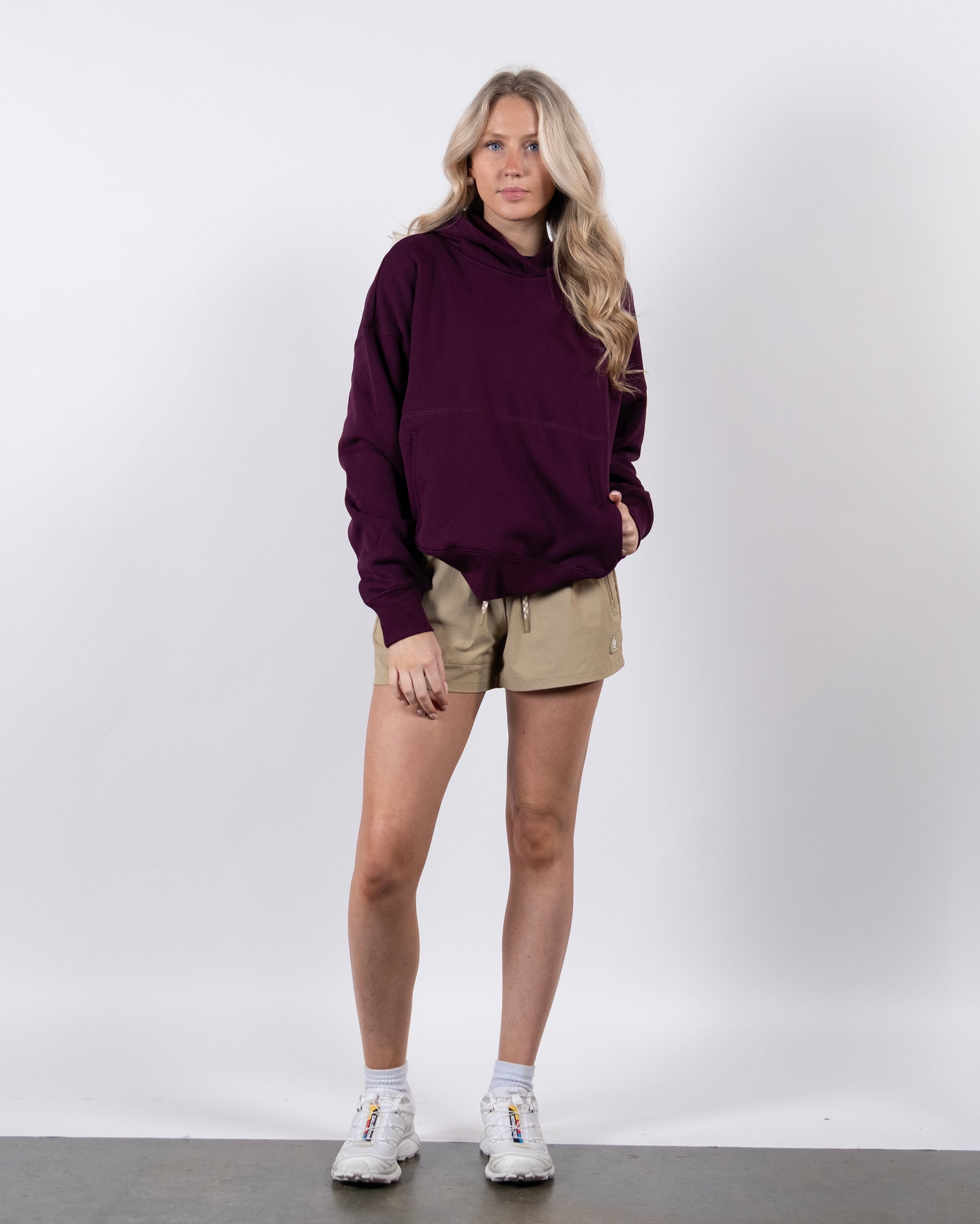 A woman with long blonde hair wears the beardedgoat Women's Overnight™ Hoodie in purple with beige shorts and white sneakers. She stands against a white background, one hand in her pocket and gazing slightly to the side.