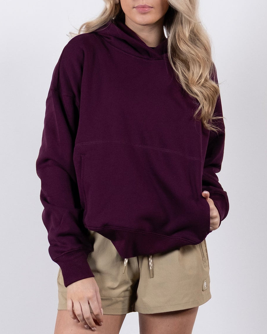 A woman with long blonde hair wears the beardedgoat Women's Overnight™ Hoodie in purple with beige shorts and white sneakers. She stands against a white background, one hand in her pocket and gazing slightly to the side.