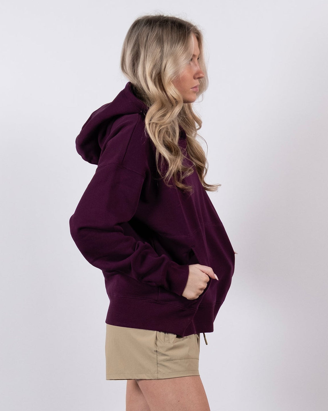 A woman with long blonde hair stands in profile against a white background, wearing a maroon beardedgoat Women's Overnight™ Hoodie, tan shorts, white socks, and white sneakers.