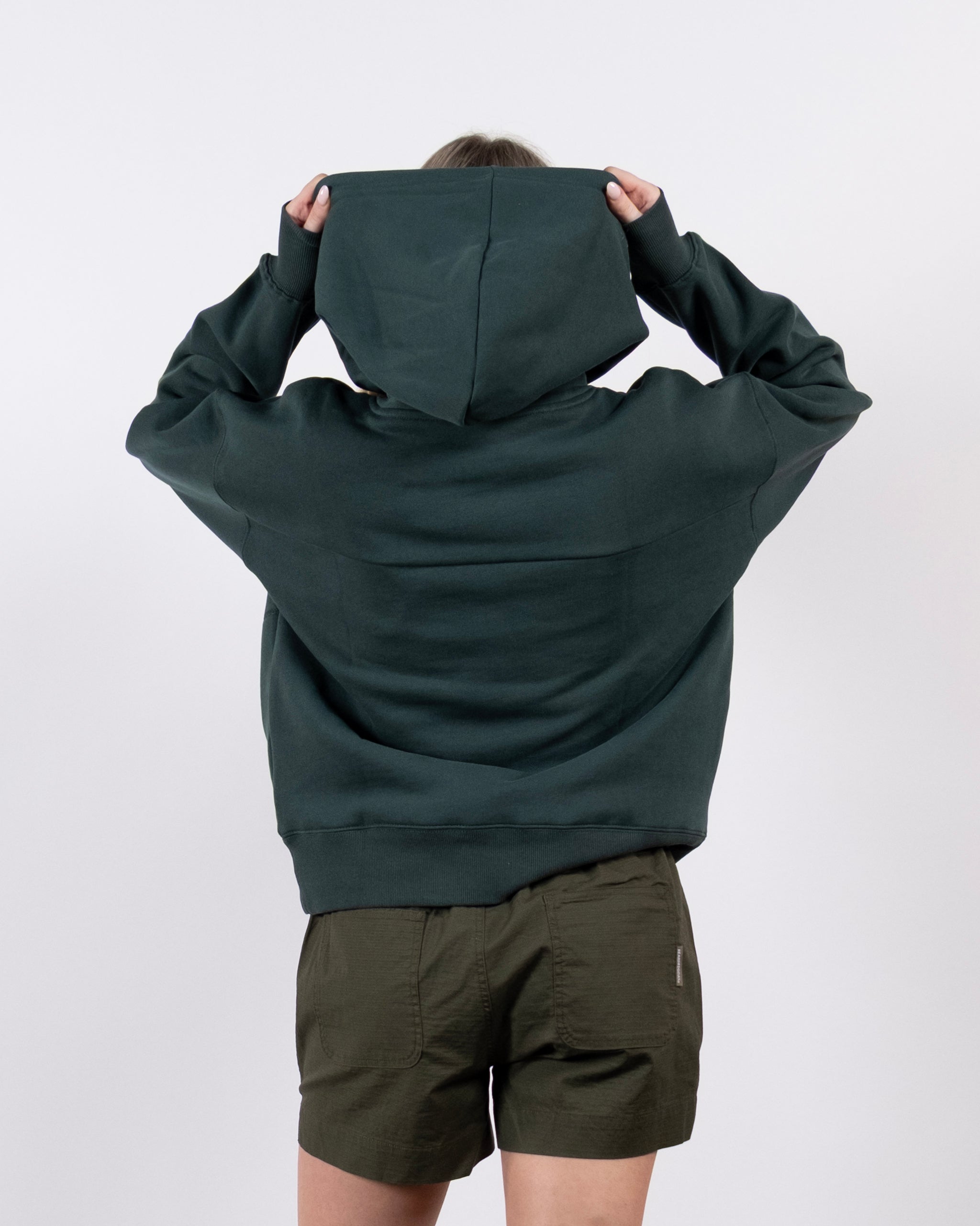 A person stands with their back to the camera, wearing a beardedgoat Women's Overnight™ Hoodie with the hood up and olive green shorts, set against a plain white background.