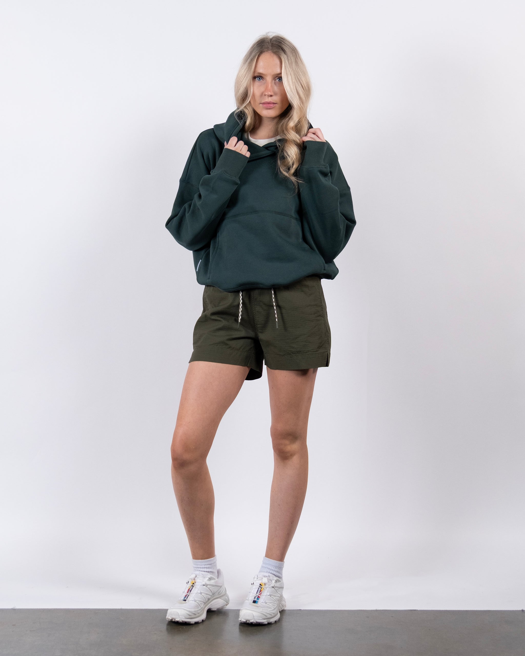 A woman with long blonde hair stands against a white background, wearing the beardedgoat Women's Overnight™ Hoodie, olive green shorts, and white sneakers. She holds the hoodie's collar with both hands and looks straight ahead.
