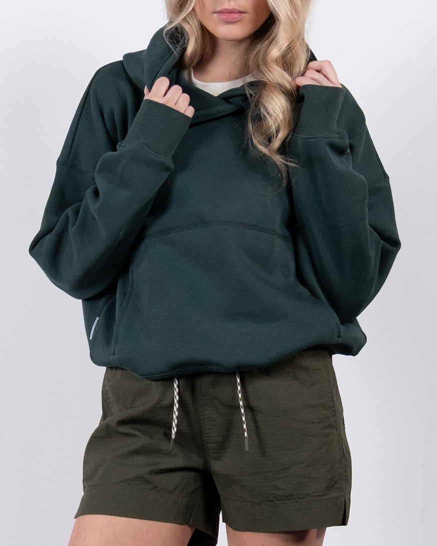 A woman with long blonde hair stands against a white background, wearing the beardedgoat Women's Overnight™ Hoodie, olive green shorts, and white sneakers. She holds the hoodie's collar with both hands and looks straight ahead.