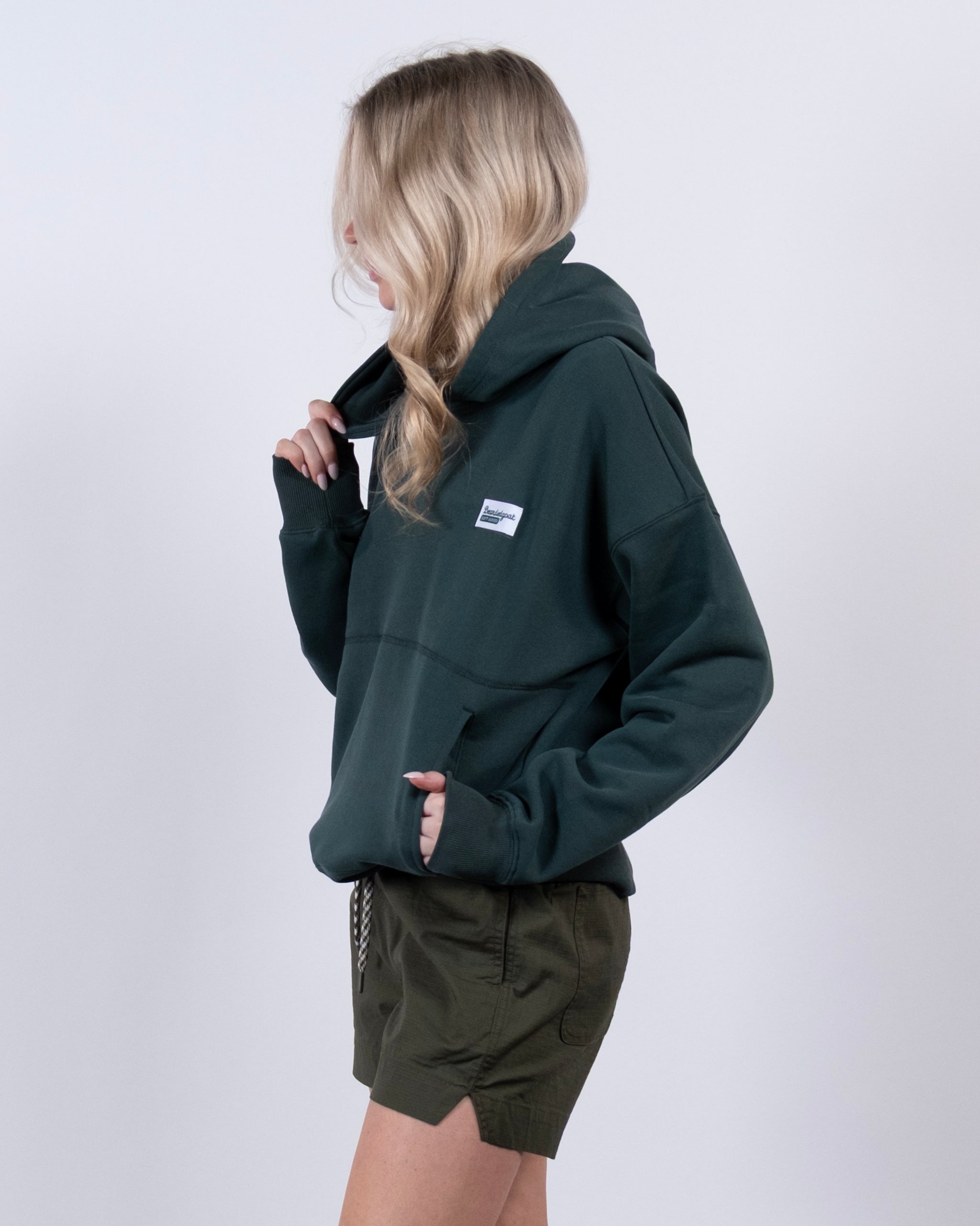 A woman with long blonde hair stands sideways against a plain light background, holding the collar of the beardedgoat Women's Overnight™ Hoodie, styled with olive green shorts for a comfortable look.