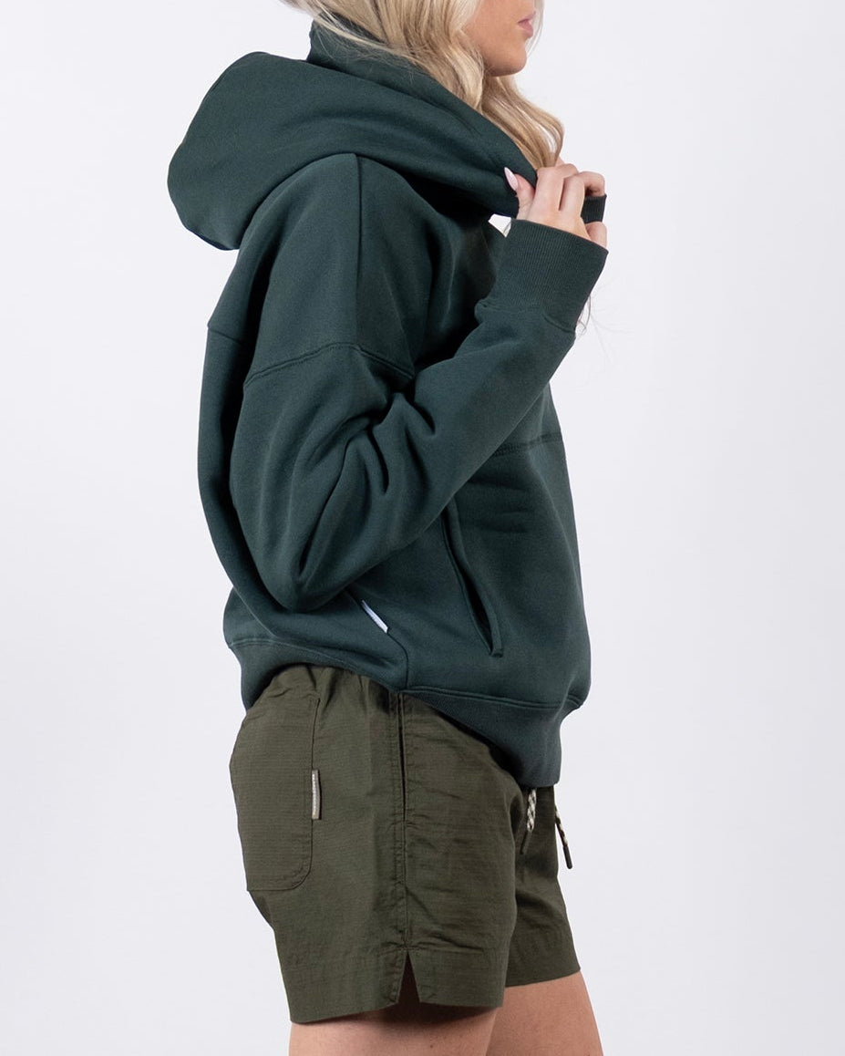A woman with light hair stands in profile against a white background, holding the beardedgoat Women's Overnight™ Hoodie close to her chin. She pairs the comfortable premium cotton fleece hoodie with olive green shorts, white socks, and sneakers.