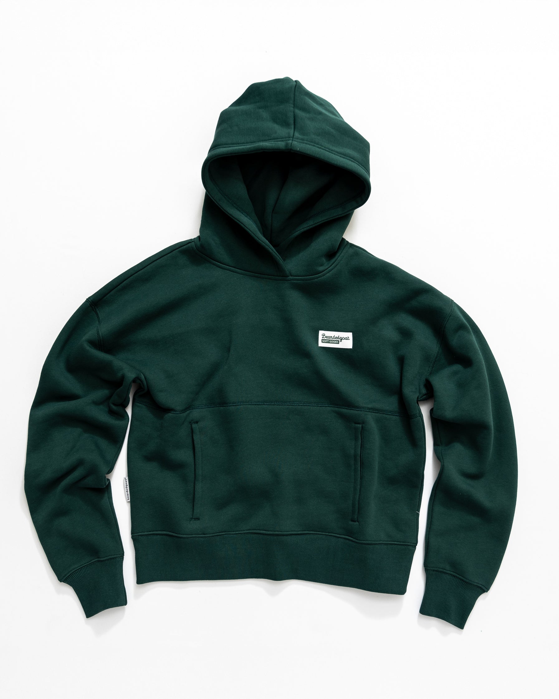 The beardedgoat Women's Overnight Hoodie in dark green is crafted from heavyweight cotton fleece, features a front pocket and a small white rectangular chest patch, and is pictured laid flat on a white surface.
