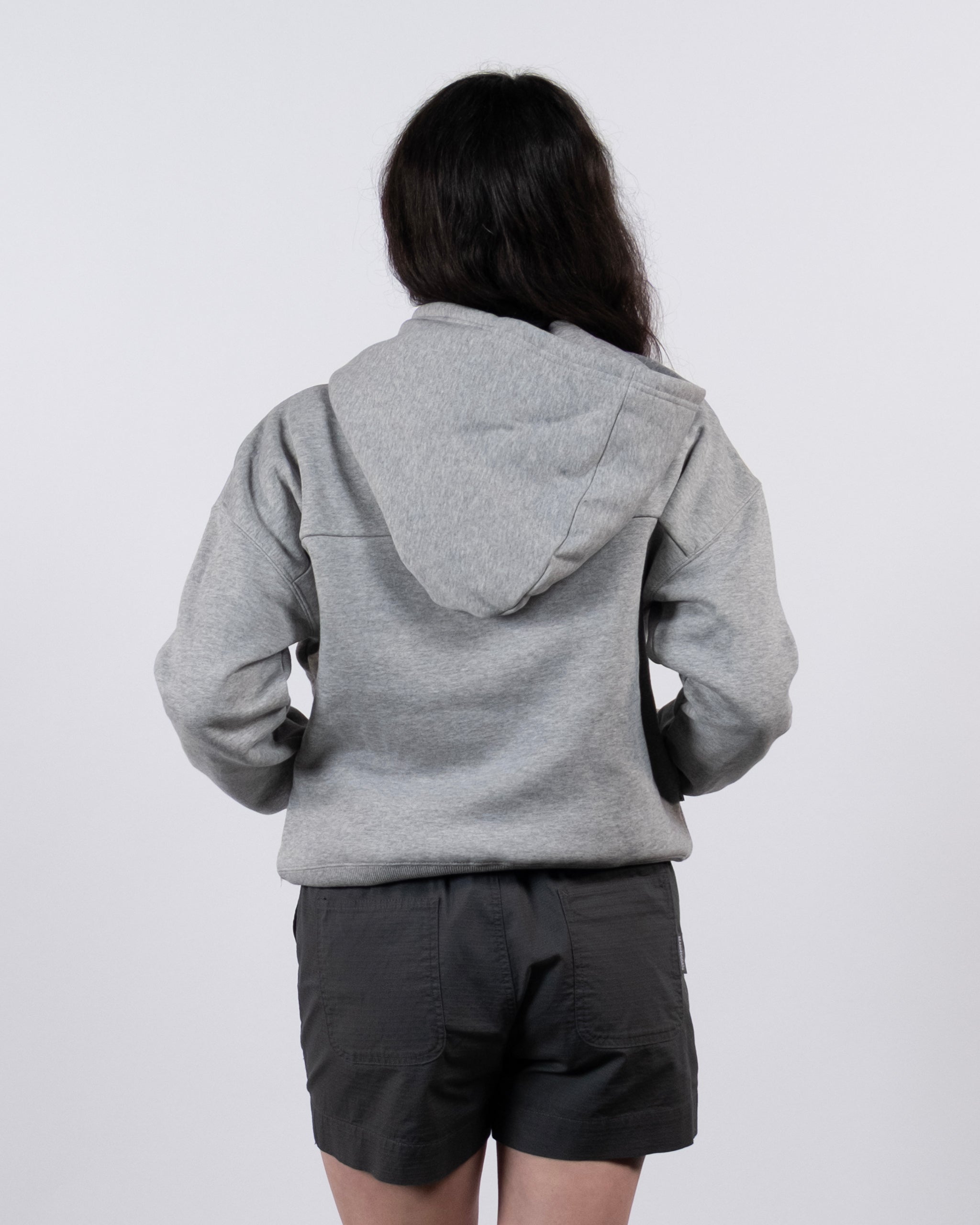 A woman with long dark hair, wearing the beardedgoat Women's Overnight™ Hoodie and dark shorts, stands facing away from the camera against a plain light background.