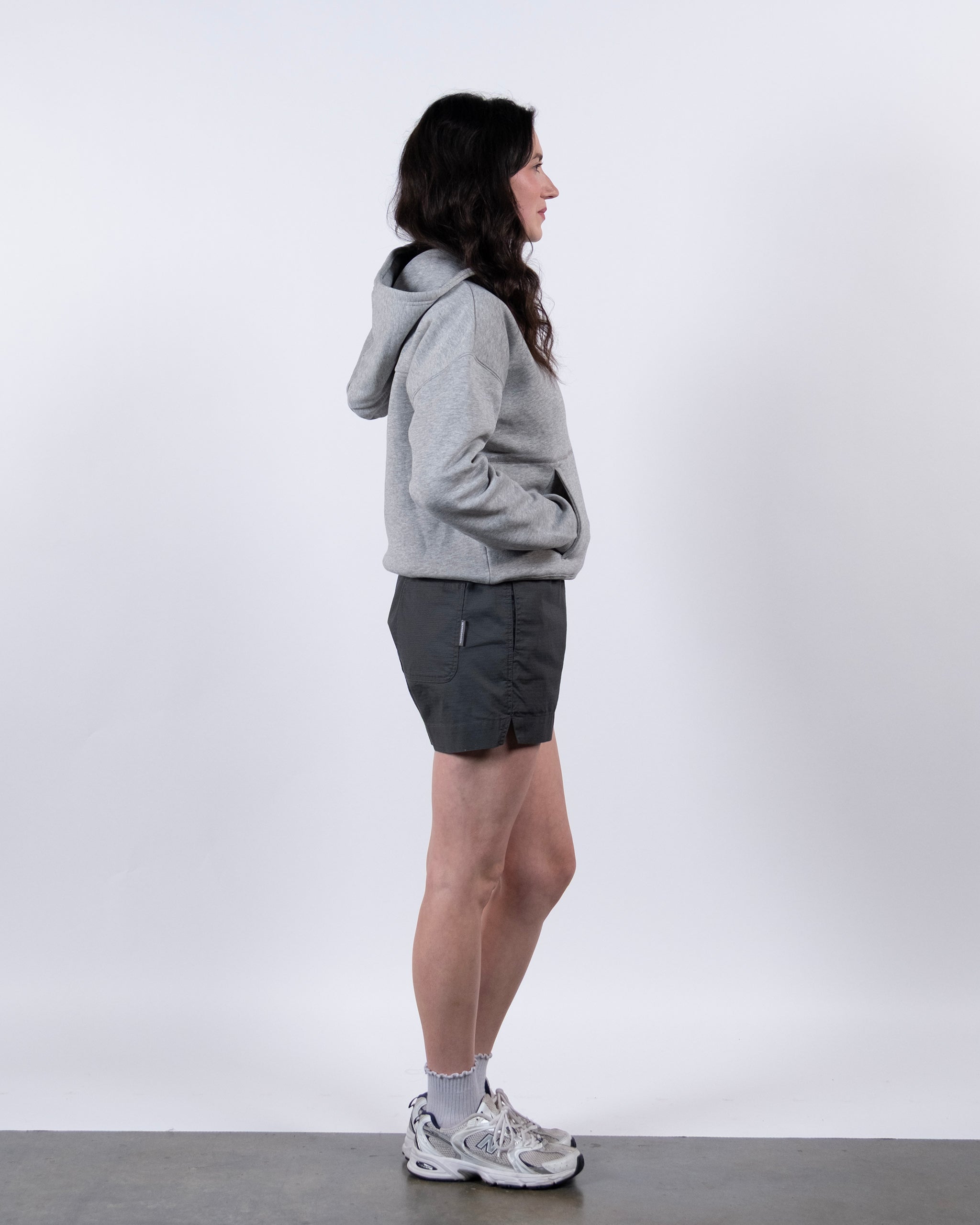 A woman with long dark hair stands in profile against a white background, wearing the beardedgoat Women's Overnight™ Hoodie, dark shorts, gray socks, and white athletic shoes with hands in her pockets.