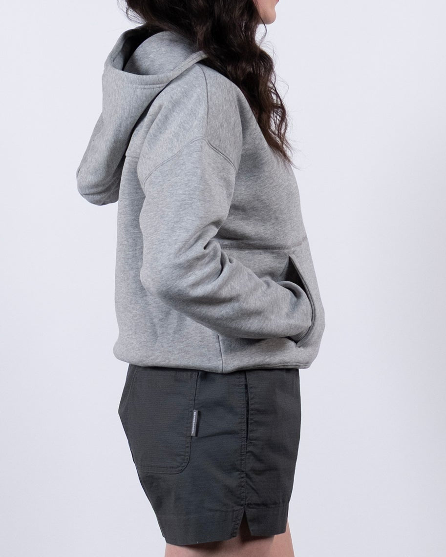 A woman with long dark hair stands in profile against a white background, wearing the beardedgoat Women's Overnight™ Hoodie, dark shorts, gray socks, and white athletic shoes with hands in her pockets.