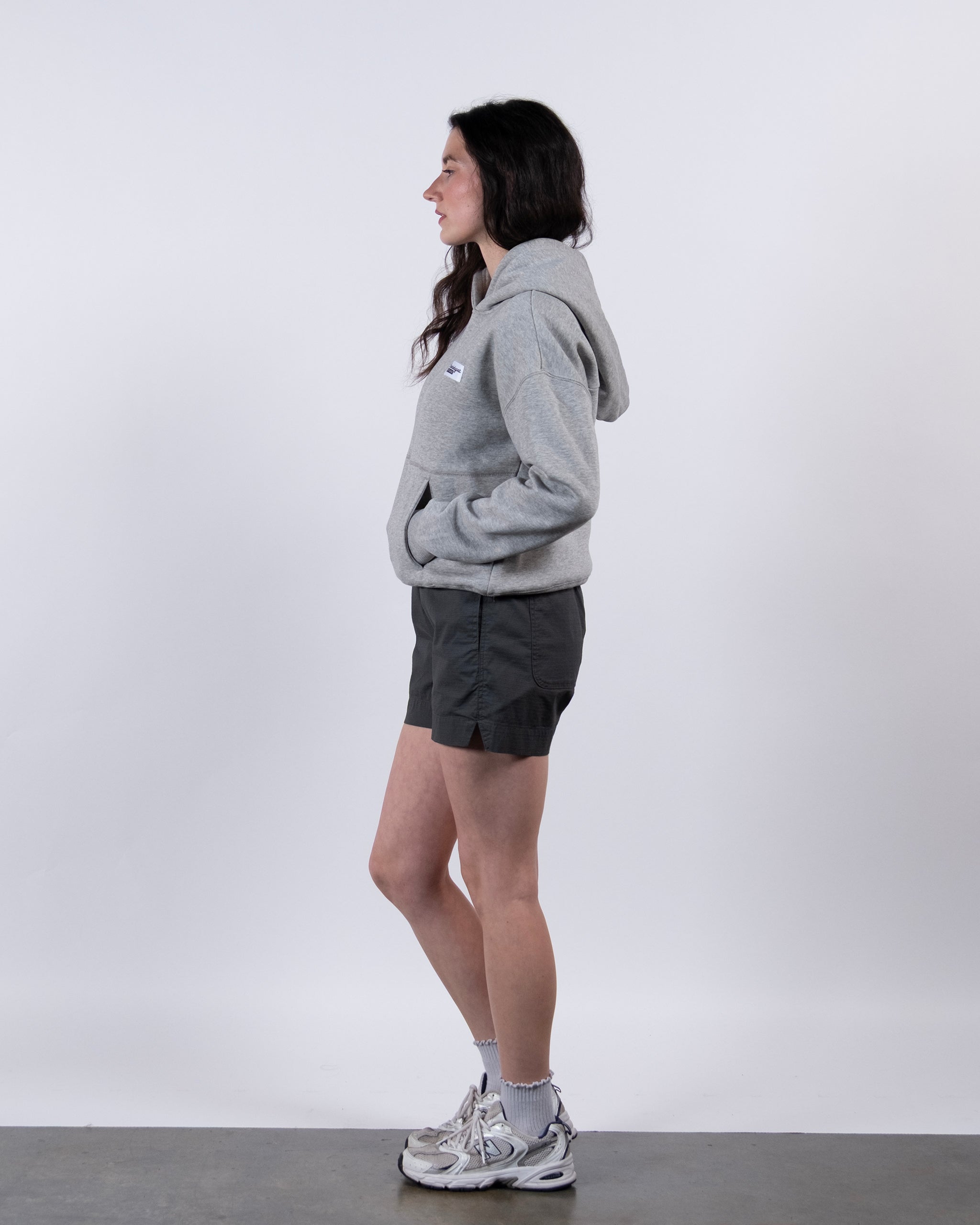 A woman stands in profile against a plain white background, wearing the beardedgoat Women's Overnight™ Hoodie in light gray, paired with dark gray shorts, gray socks, and white sneakers, with her hands tucked into the hoodie pocket.