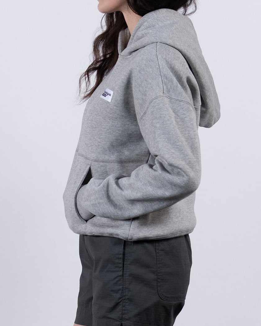 A woman stands in profile against a plain white background, wearing the beardedgoat Women's Overnight™ Hoodie in light gray, paired with dark gray shorts, gray socks, and white sneakers, with her hands tucked into the hoodie pocket.