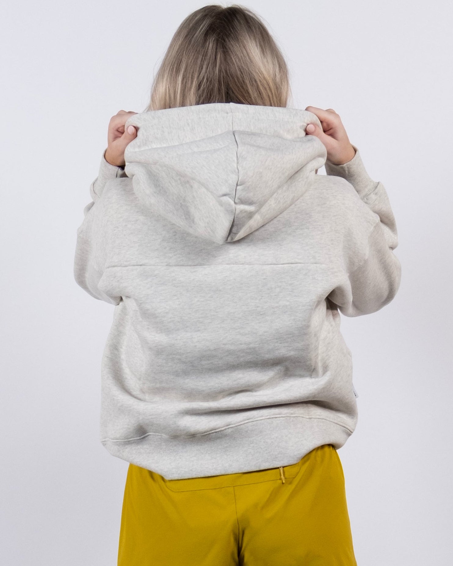 A person with blonde hair, back to camera, wears a light gray beardedgoat Women's Overnight™ Hoodie and yellow shorts against a plain white background, holding up the hood with both hands.