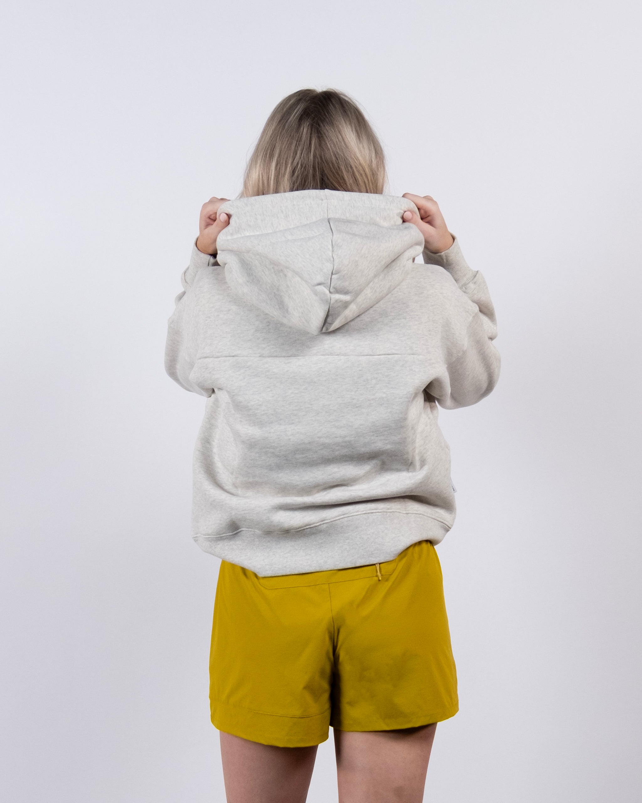 A person with blonde hair, back to camera, wears a light gray beardedgoat Women's Overnight™ Hoodie and yellow shorts against a plain white background, holding up the hood with both hands.