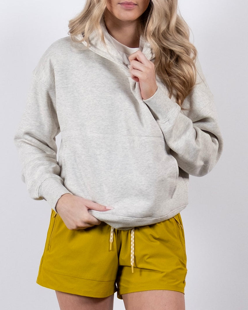 A woman with long blonde hair wears the beardedgoat Women's Overnight™ Hoodie in light gray. She stands against a plain white background, facing the camera with one hand near her neck, paired with mustard shorts, white socks, and sneakers.