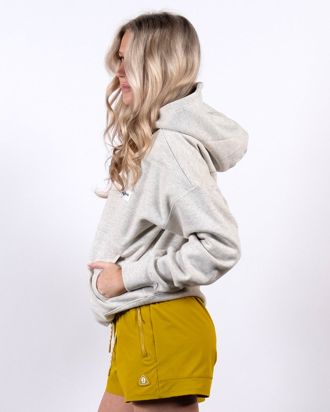 A woman stands in profile against a white background, wearing a light gray beardedgoat Women's Overnight™ Hoodie, yellow shorts, white ankle socks, and white sneakers. Her hands are in the hoodie pocket and her long blonde hair is down.