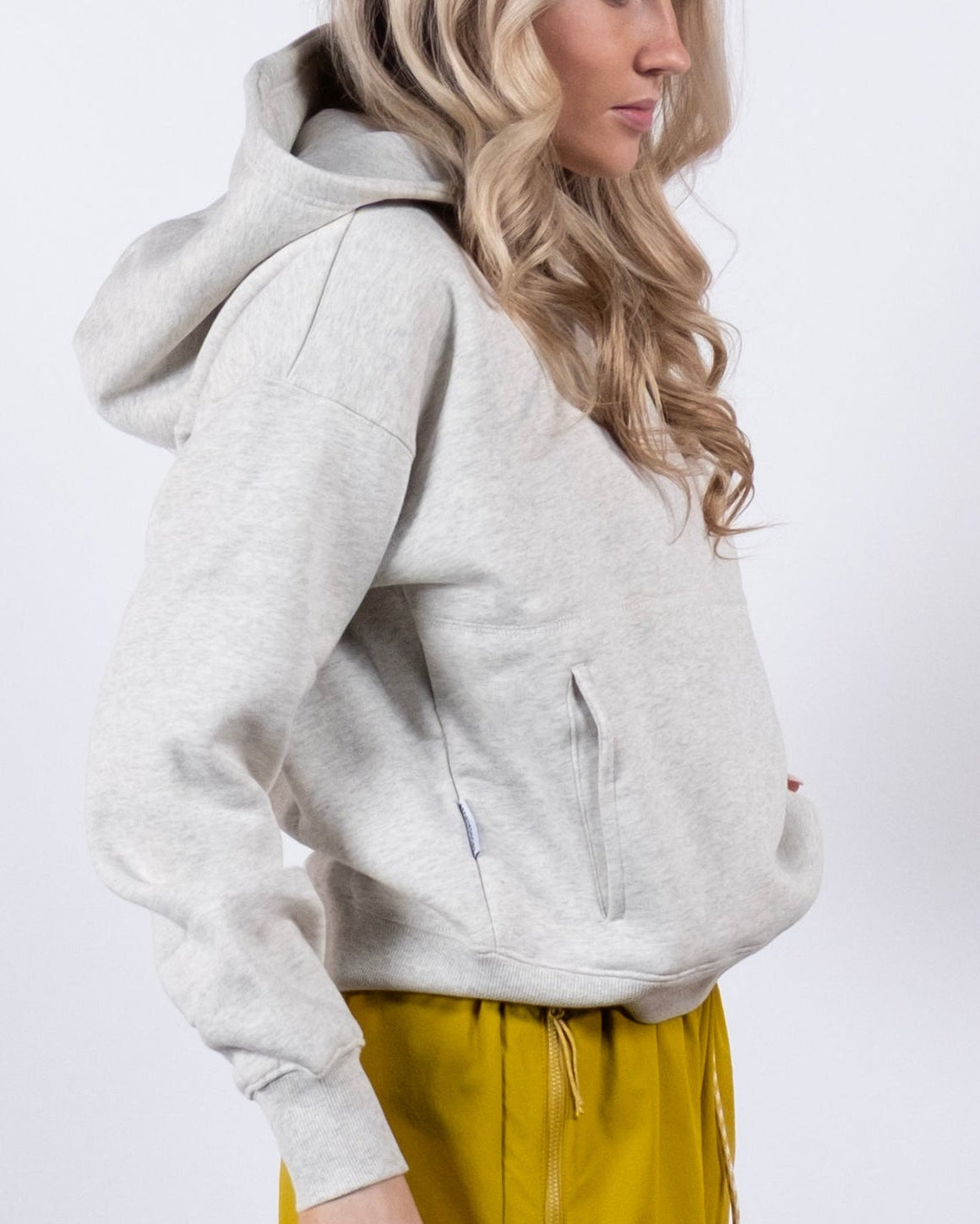 A woman with long, wavy blonde hair wears the beardedgoat Women's Overnight™ Hoodie and yellow shorts, standing sideways against a plain white background.