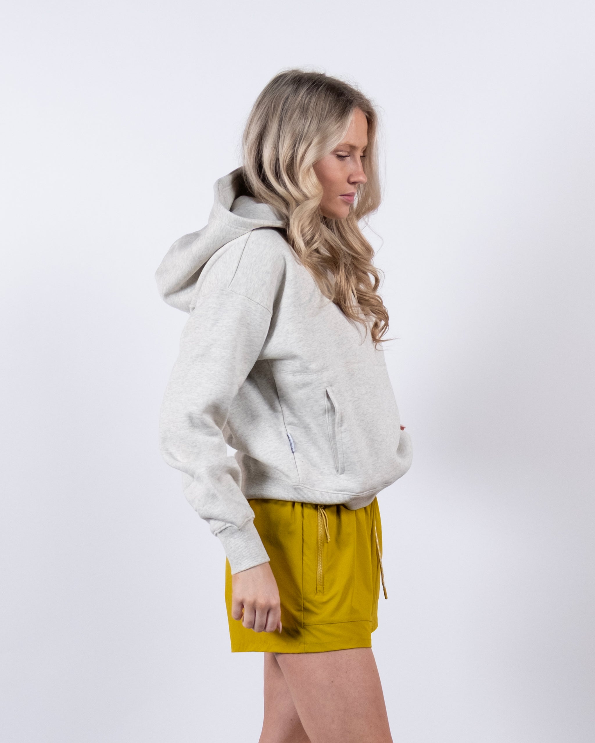 A woman with long, wavy blonde hair wears the beardedgoat Women's Overnight™ Hoodie and yellow shorts, standing sideways against a plain white background.