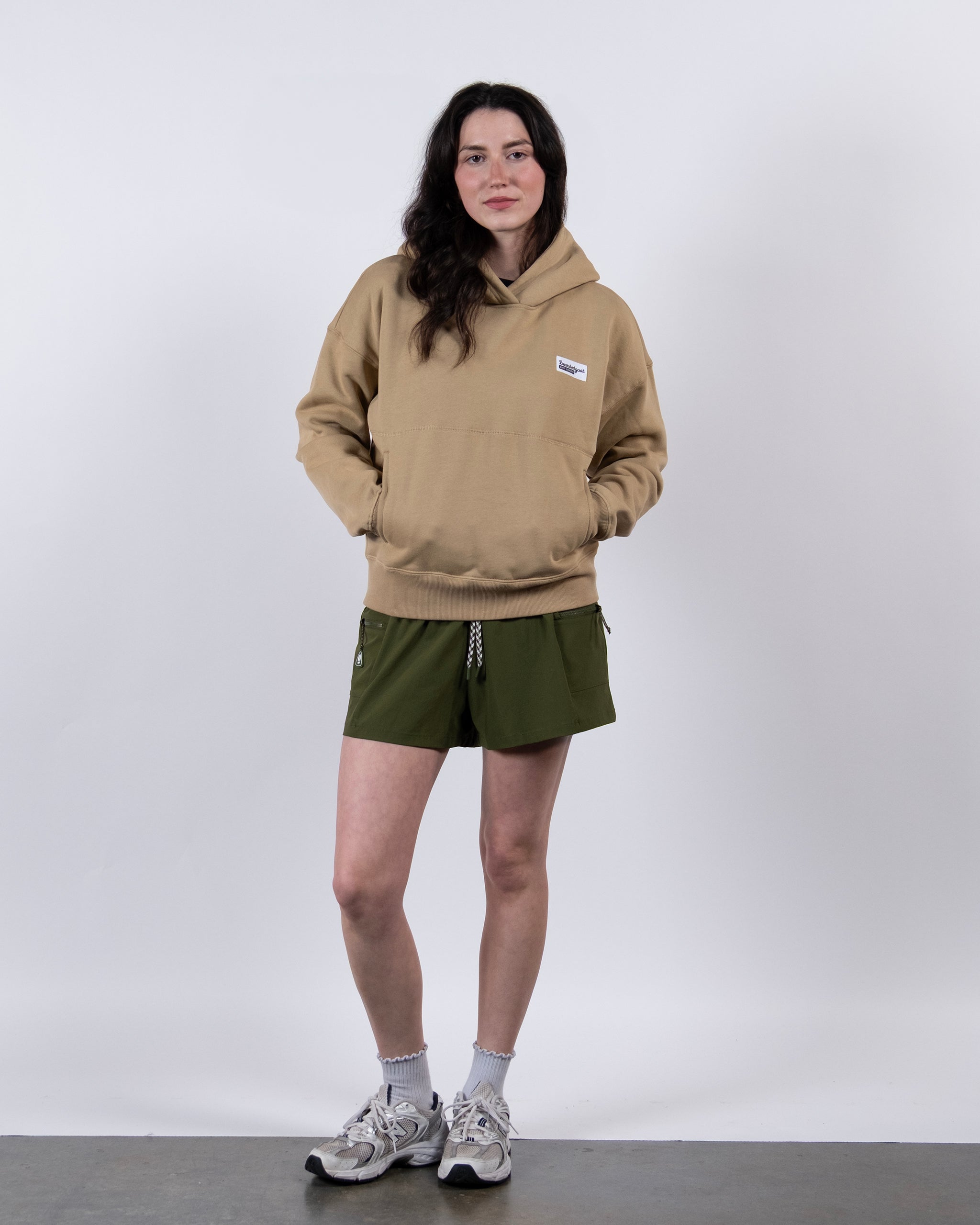 A woman stands against a white background, wearing the beardedgoat Women's Overnight™ Hoodie, styled with olive green shorts, white socks, and gray sneakers. Her hands are in the hoodie pocket as she looks at the camera with a slight smile.