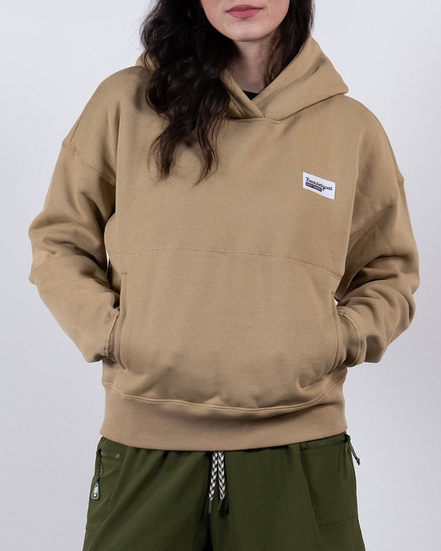 A woman stands against a white background, wearing the beardedgoat Women's Overnight™ Hoodie, styled with olive green shorts, white socks, and gray sneakers. Her hands are in the hoodie pocket as she looks at the camera with a slight smile.