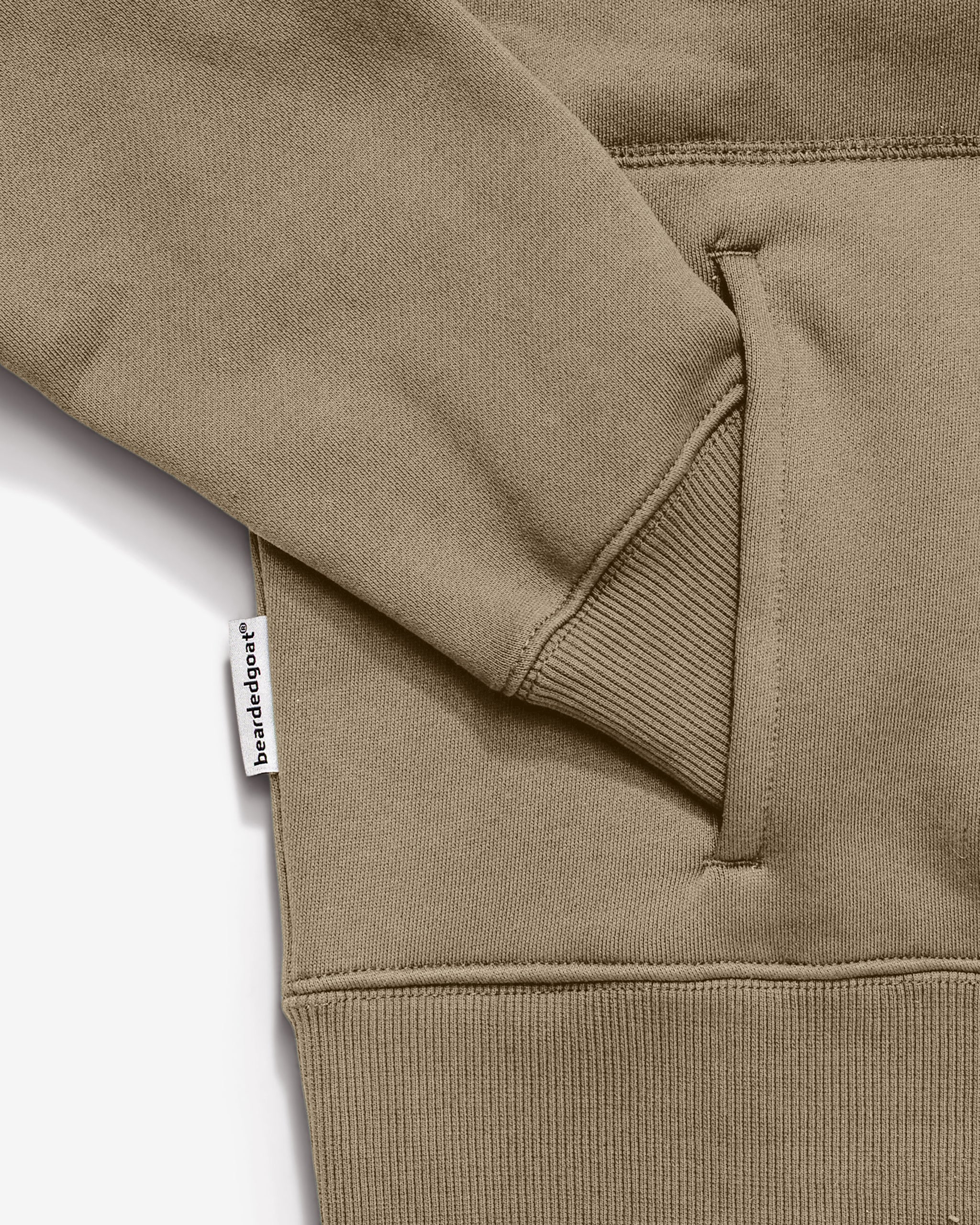 Close-up of a Women's Overnight™ Hoodie by beardedgoat in tan or khaki, showing the ribbed cuffs and hem made from premium cotton fleece and a small white brandedspot tag on the side seam.